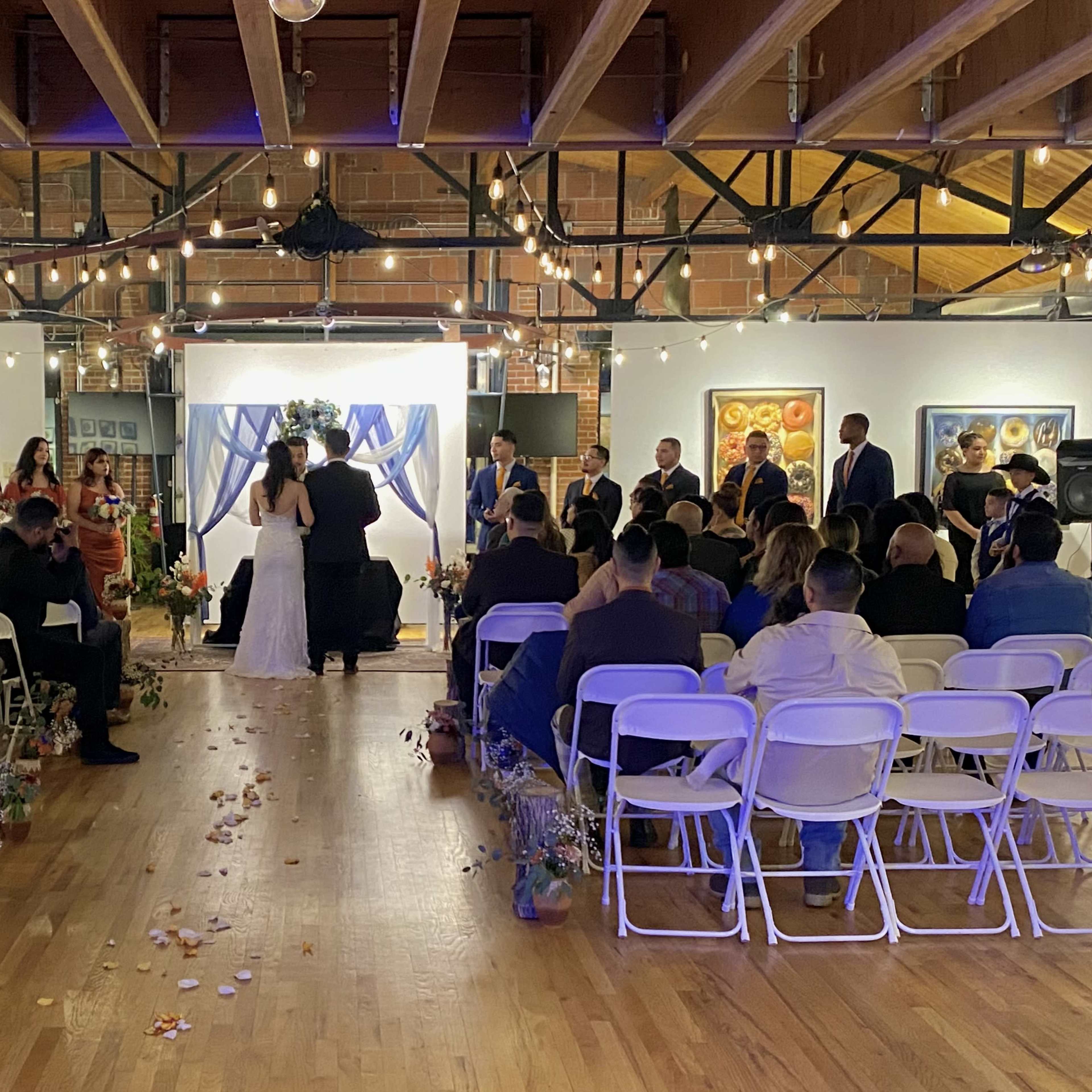 A wedding ceremony is taking place indoors, with guests seated on white chairs and a decorated stage featuring a couple exchanging vows.