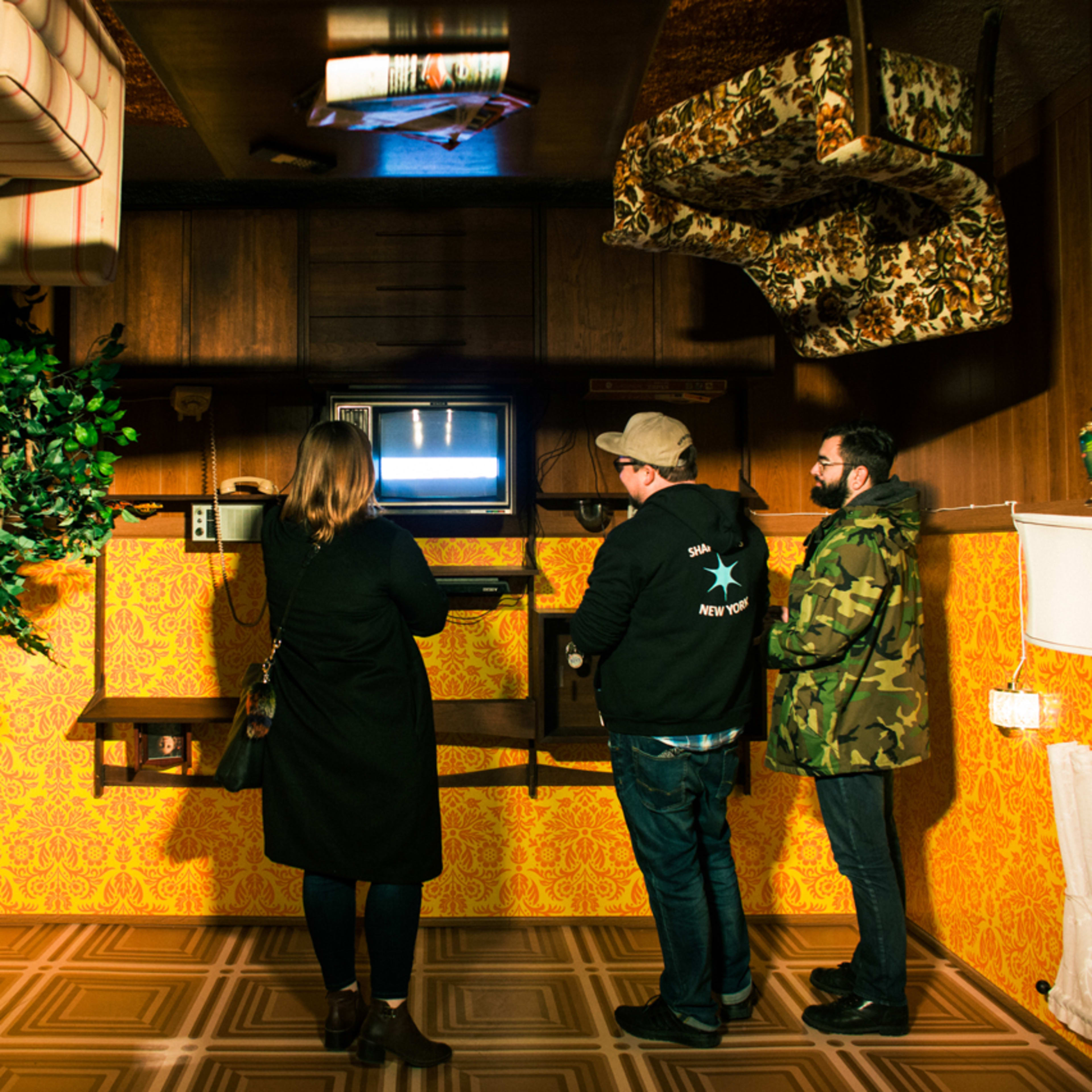Three people stand in a room with upside-down furniture, watching a television screen.