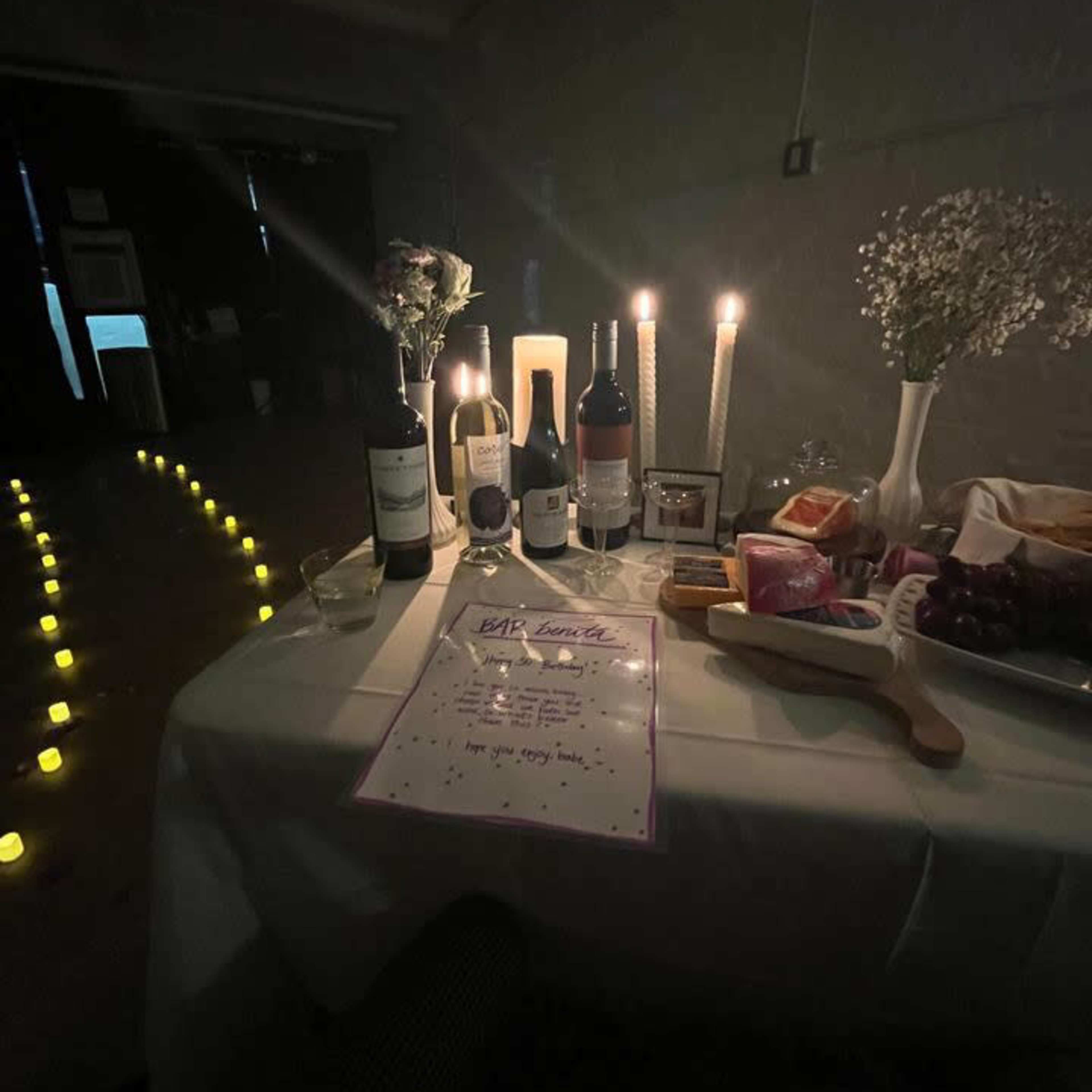 A table is set with wine bottles, candles, and a menu, surrounded by glowing candles leading to a darkened room.