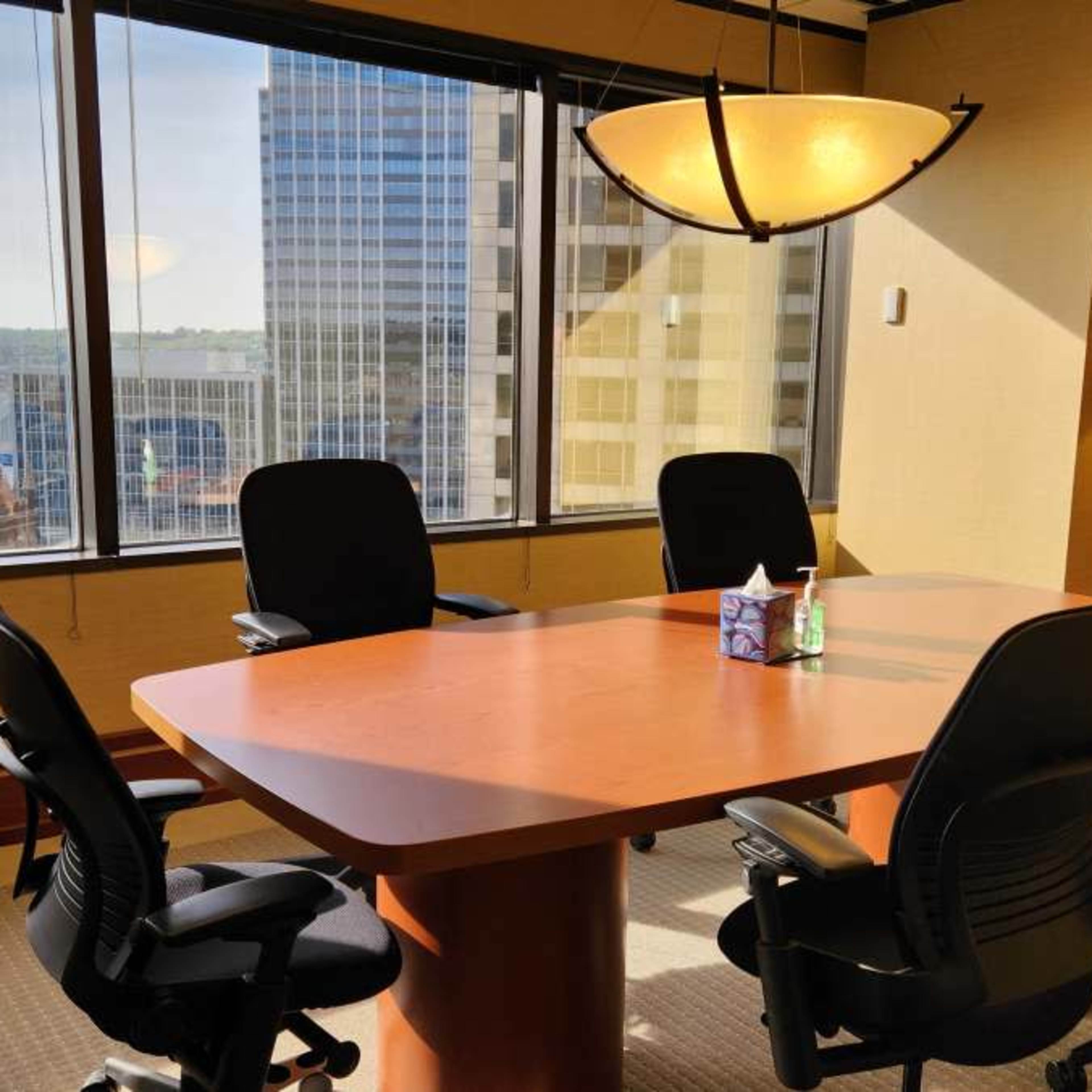 A conference room features a large wooden table surrounded by four chairs, with a view of city buildings and a river visible through a large window.