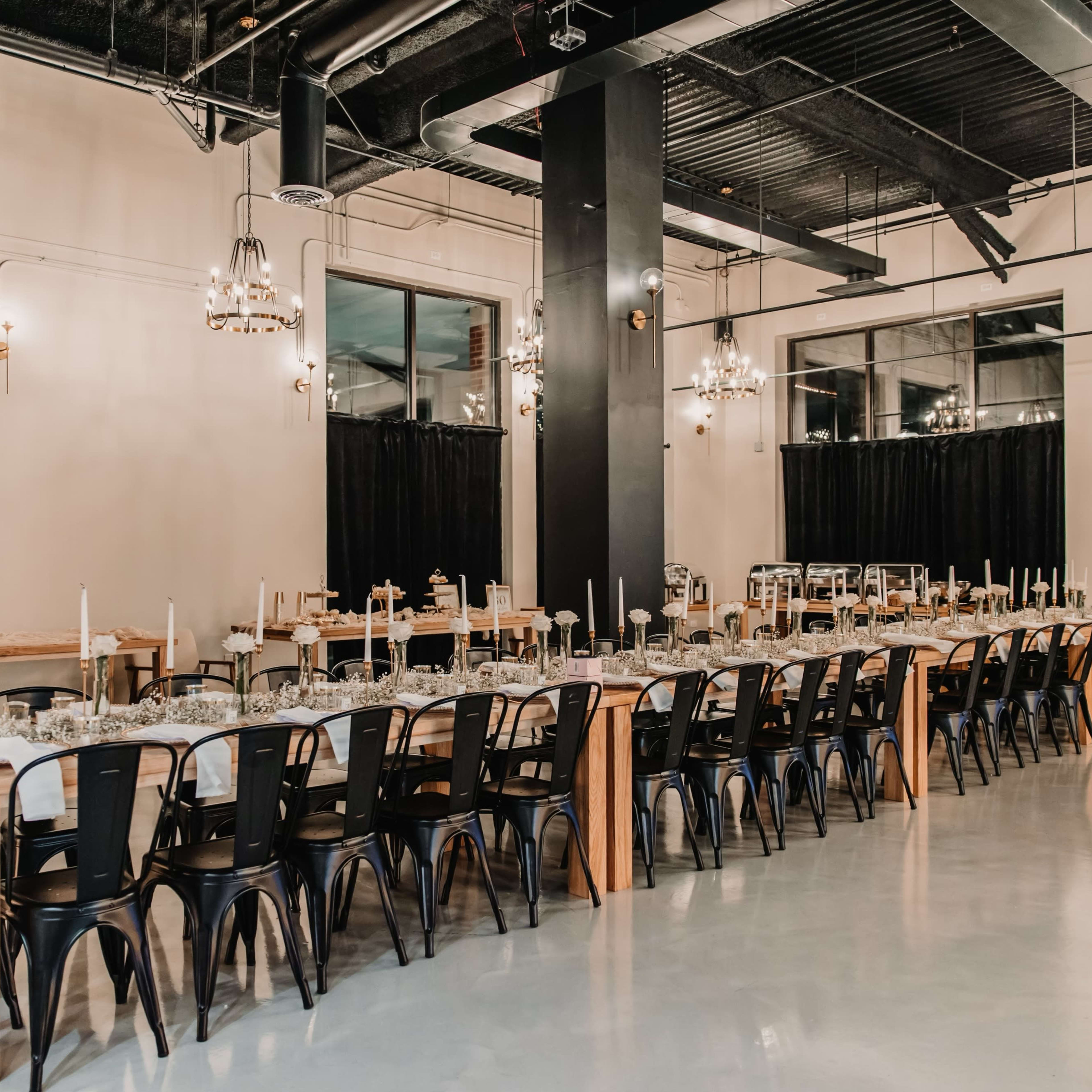 A long wooden dining table is set with candles and floral arrangements in a modern event space with industrial lighting and dark curtains.