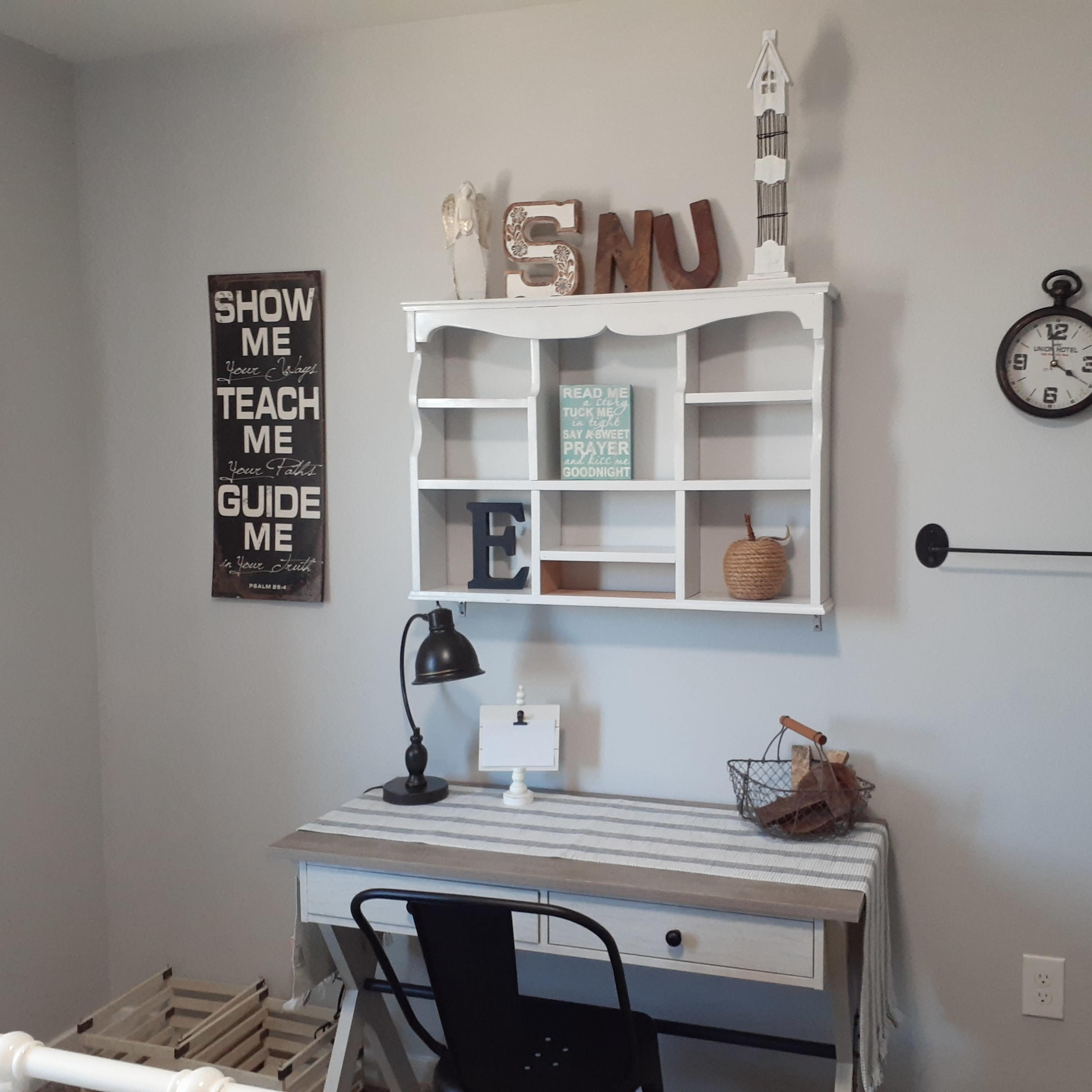A small, organized workspace with a white desk, black chair, and decorative wall shelves displaying various items and a clock.