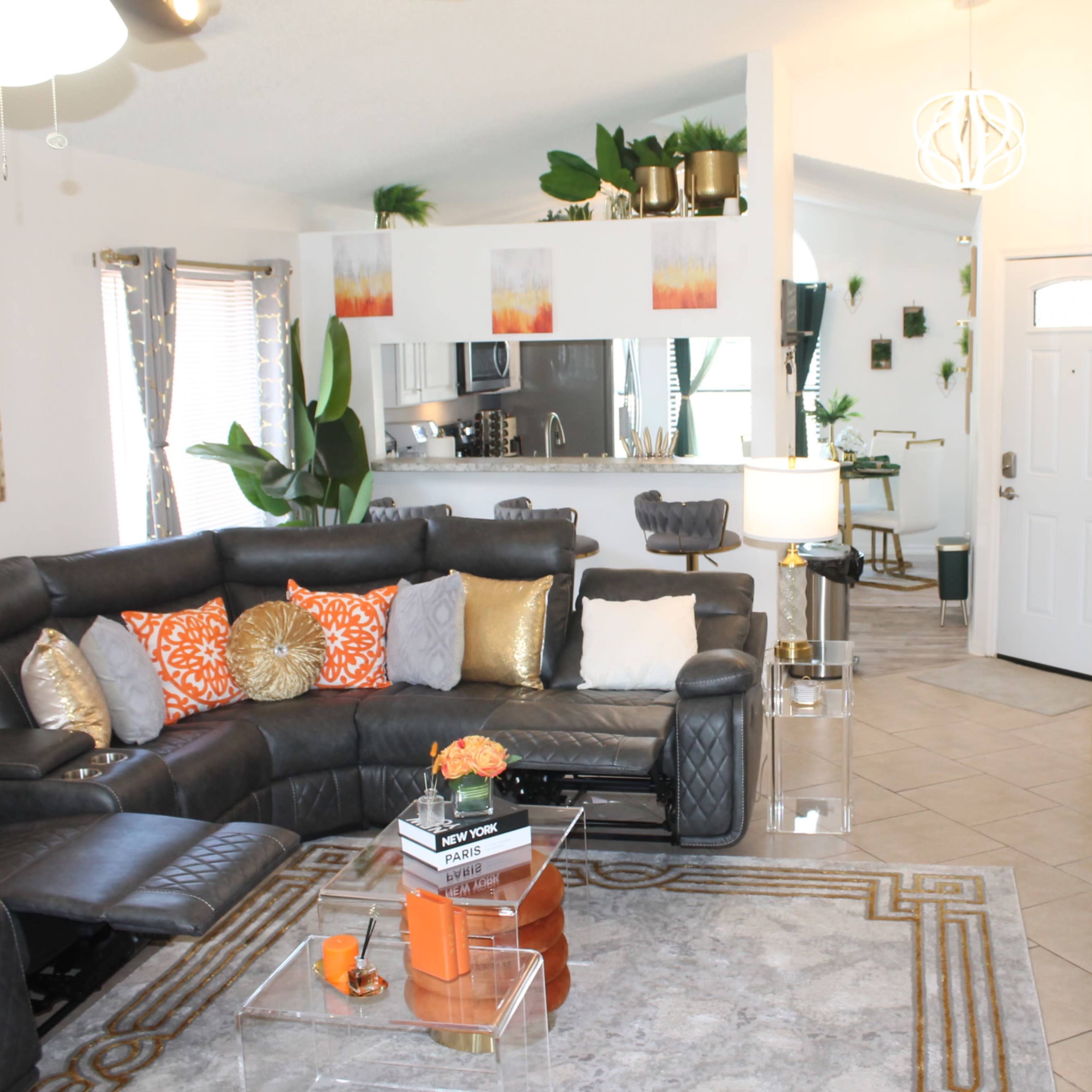 A modern living room with a sectional sofa, colorful cushions, a glass coffee table, and a doorway leading to a kitchen area adorned with plants and decorative accents.