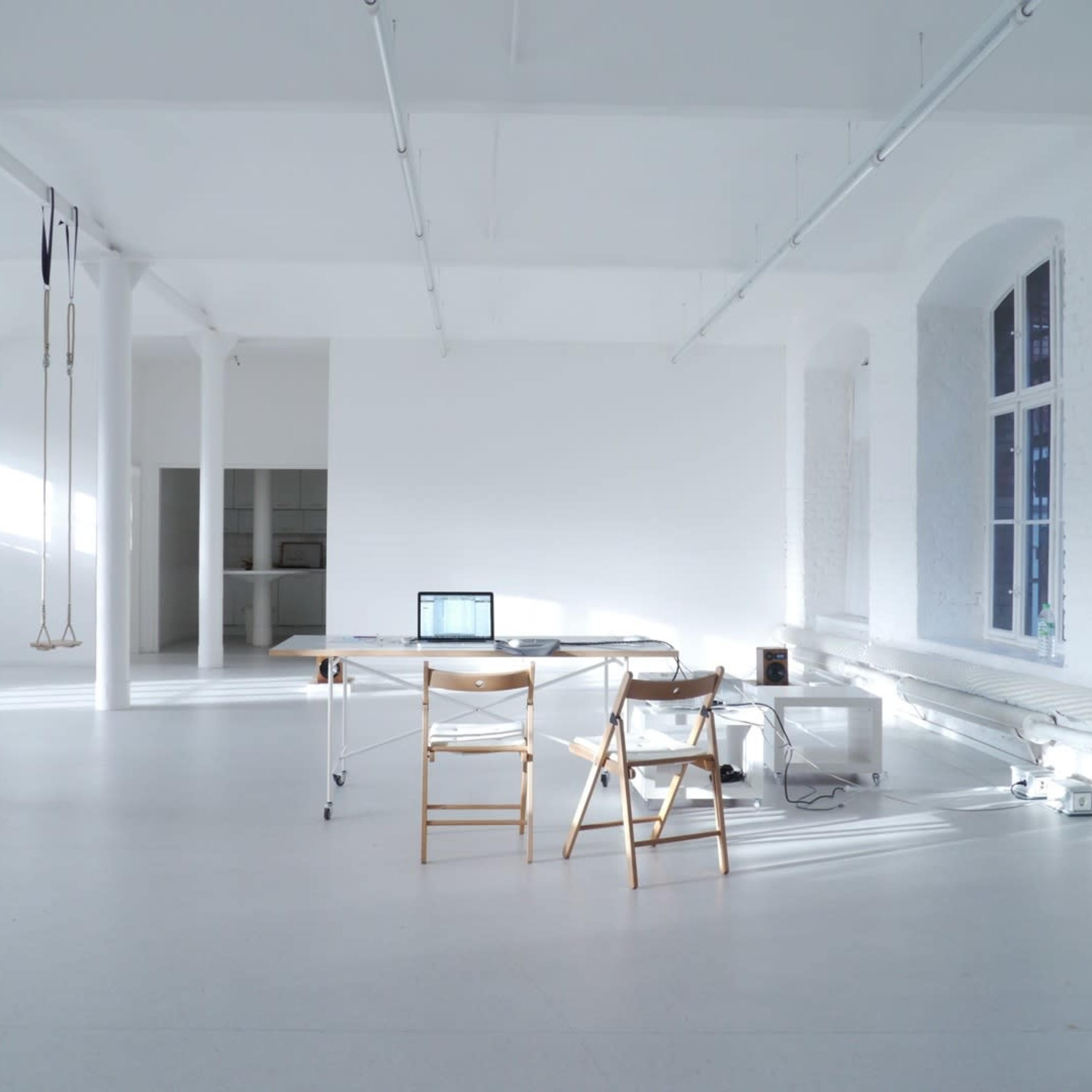 A large, minimalistic room with white walls and floors features a simple desk, two chairs, and a laptop, illuminated by natural light from windows.