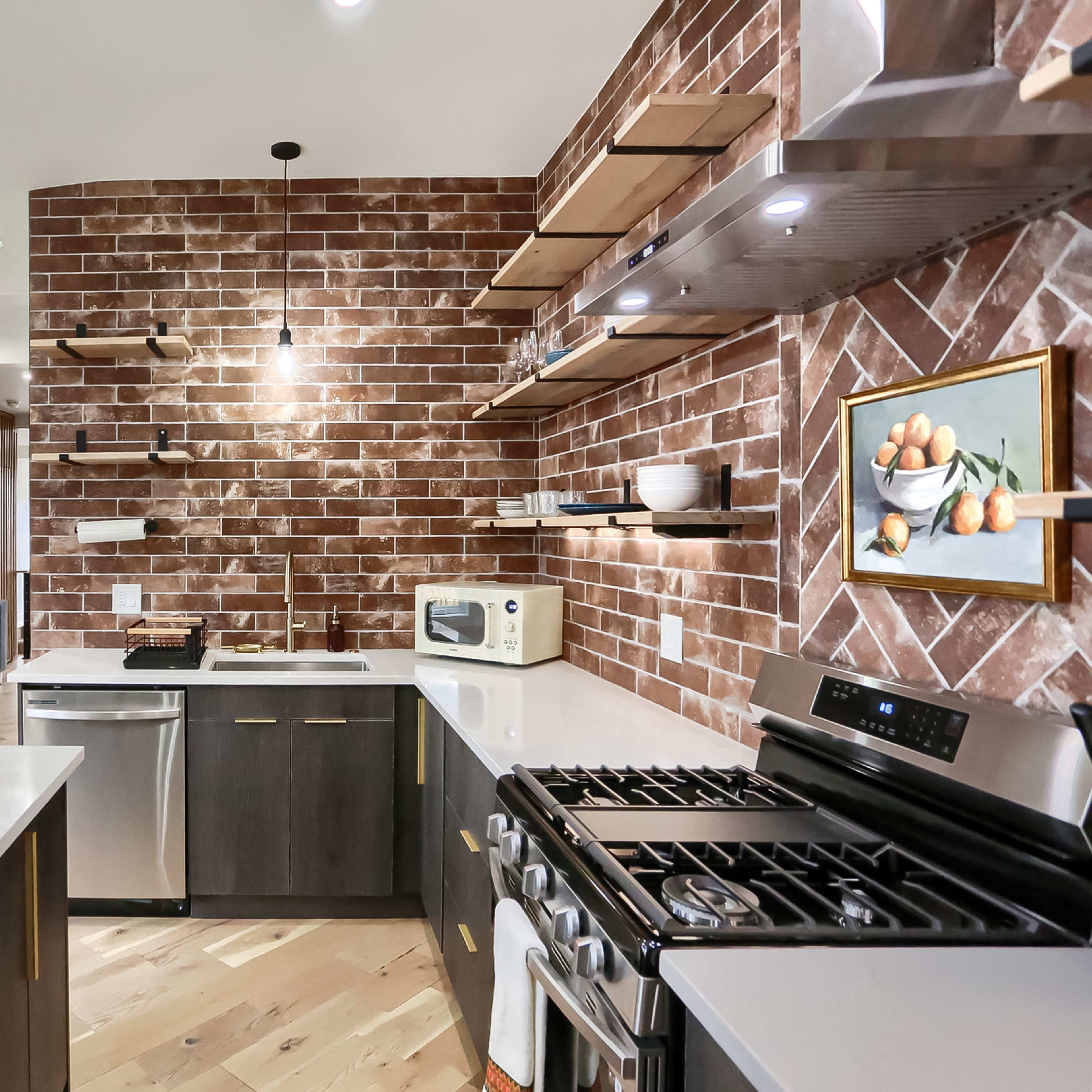 A modern kitchen features a brick accent wall, stainless steel appliances, and stylish open shelves displaying dishware.