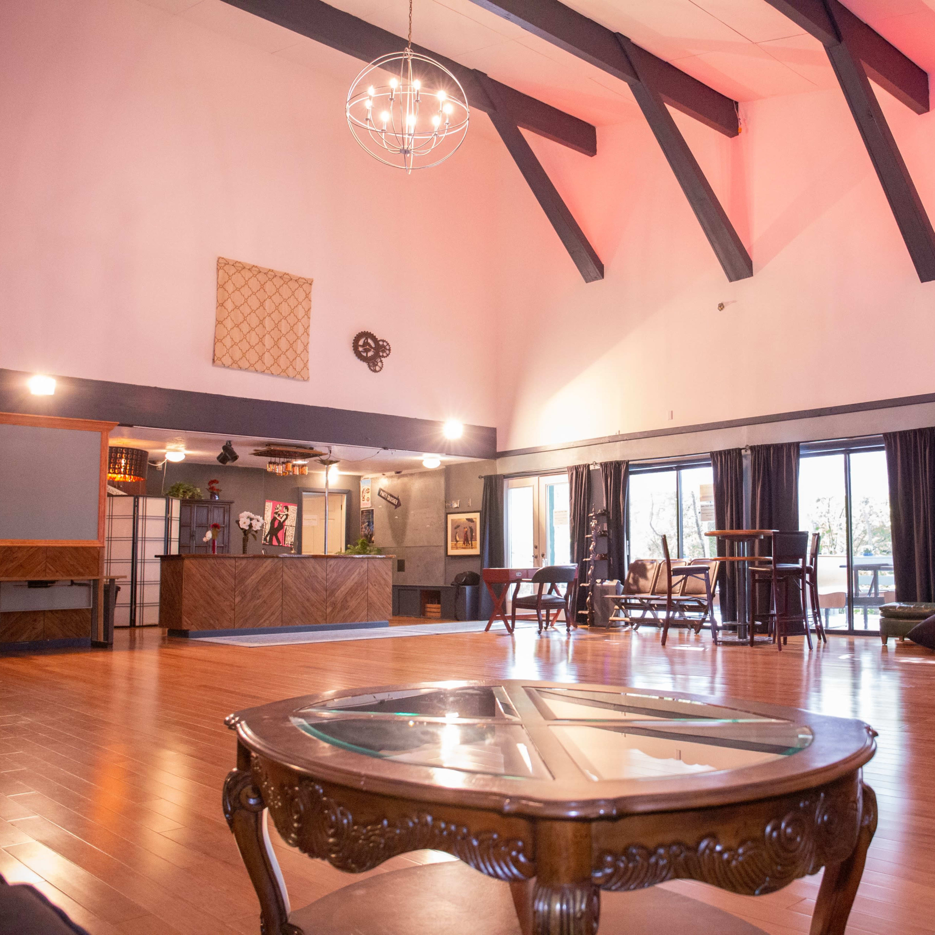 A spacious room with high ceilings features wooden flooring, large windows, and various seating arrangements.