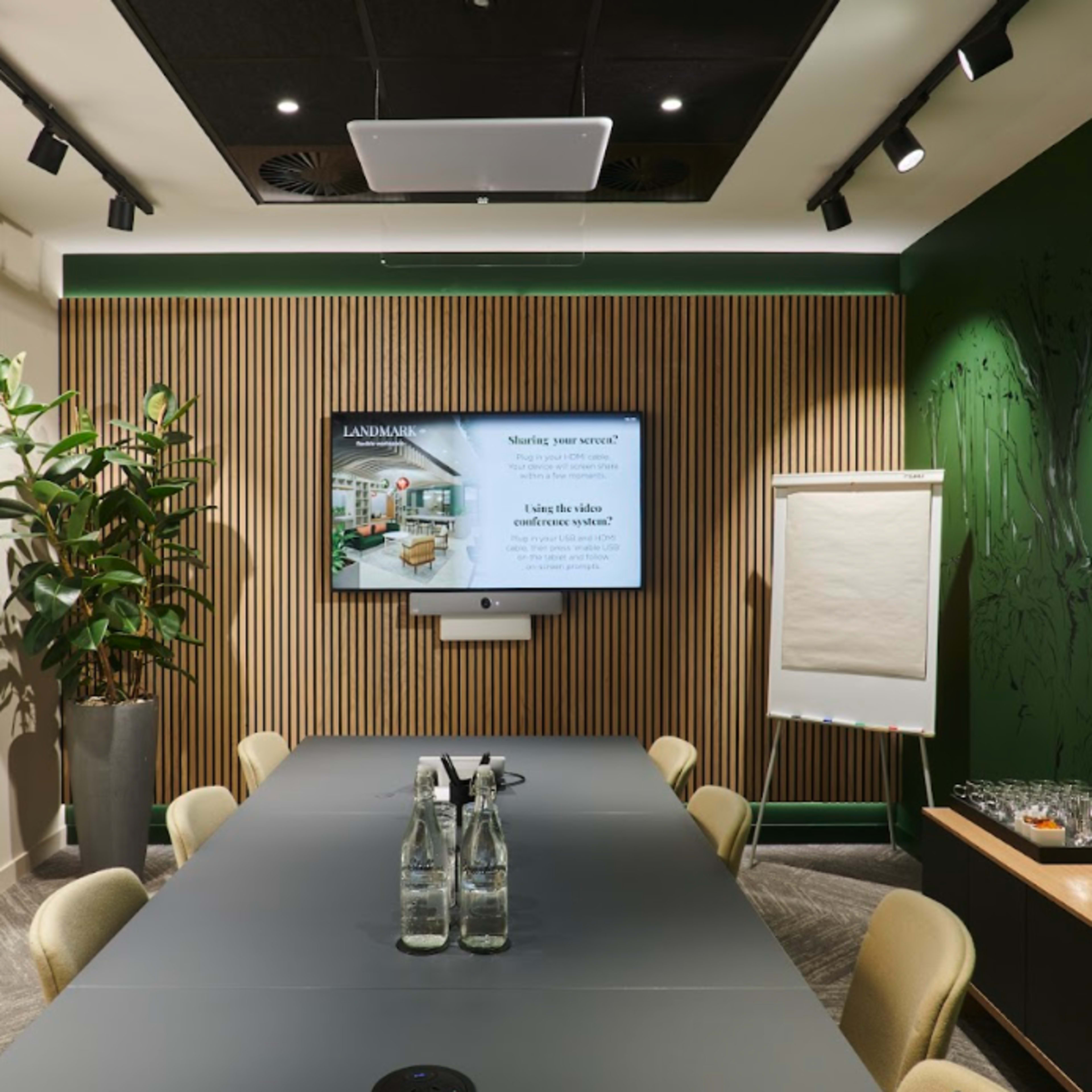 A modern meeting room features a long table, a large screen displaying a presentation, and plants against a wood-paneled wall.