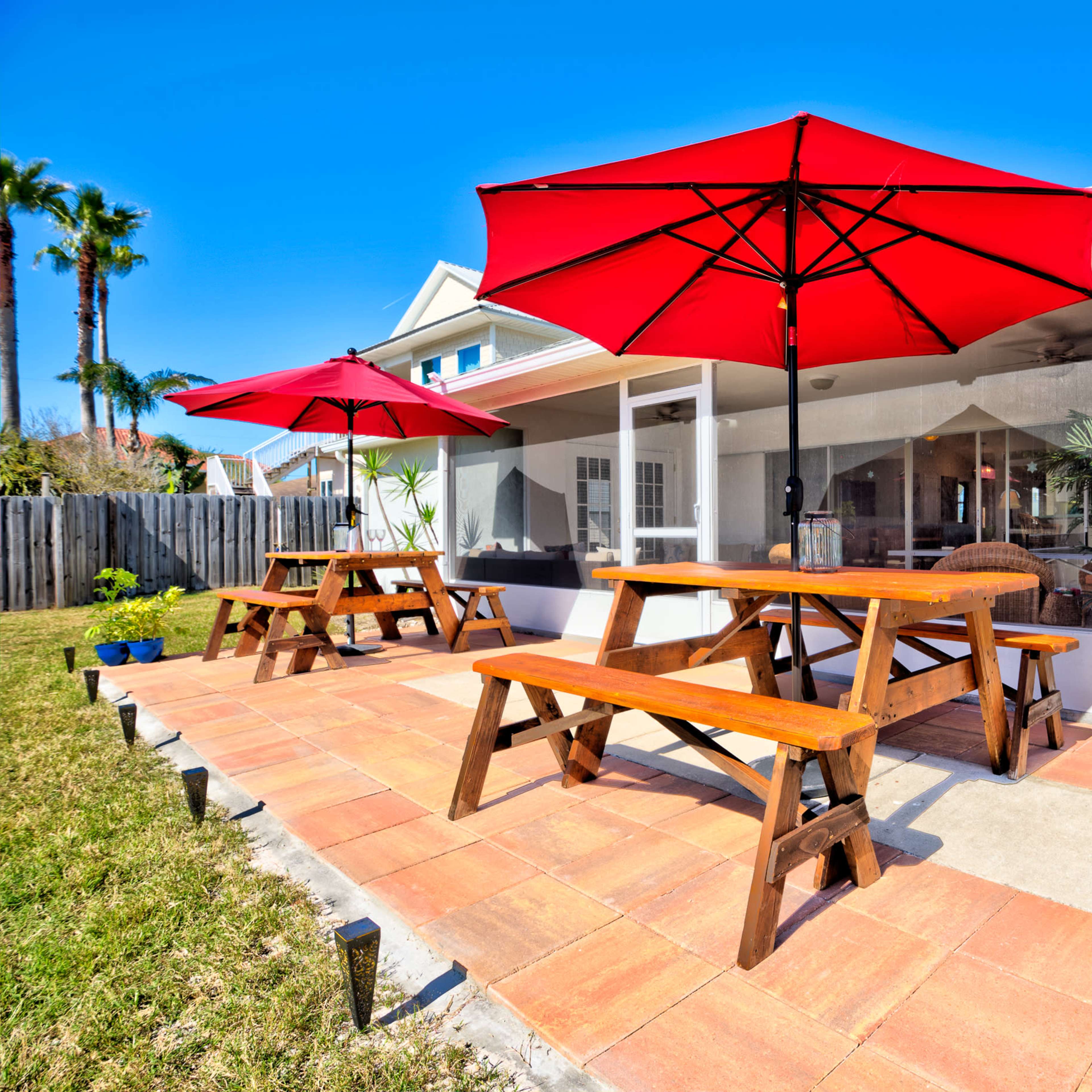A patio area with two red umbrellas over wooden picnic tables and a grill, surrounded by grass and potted plants.