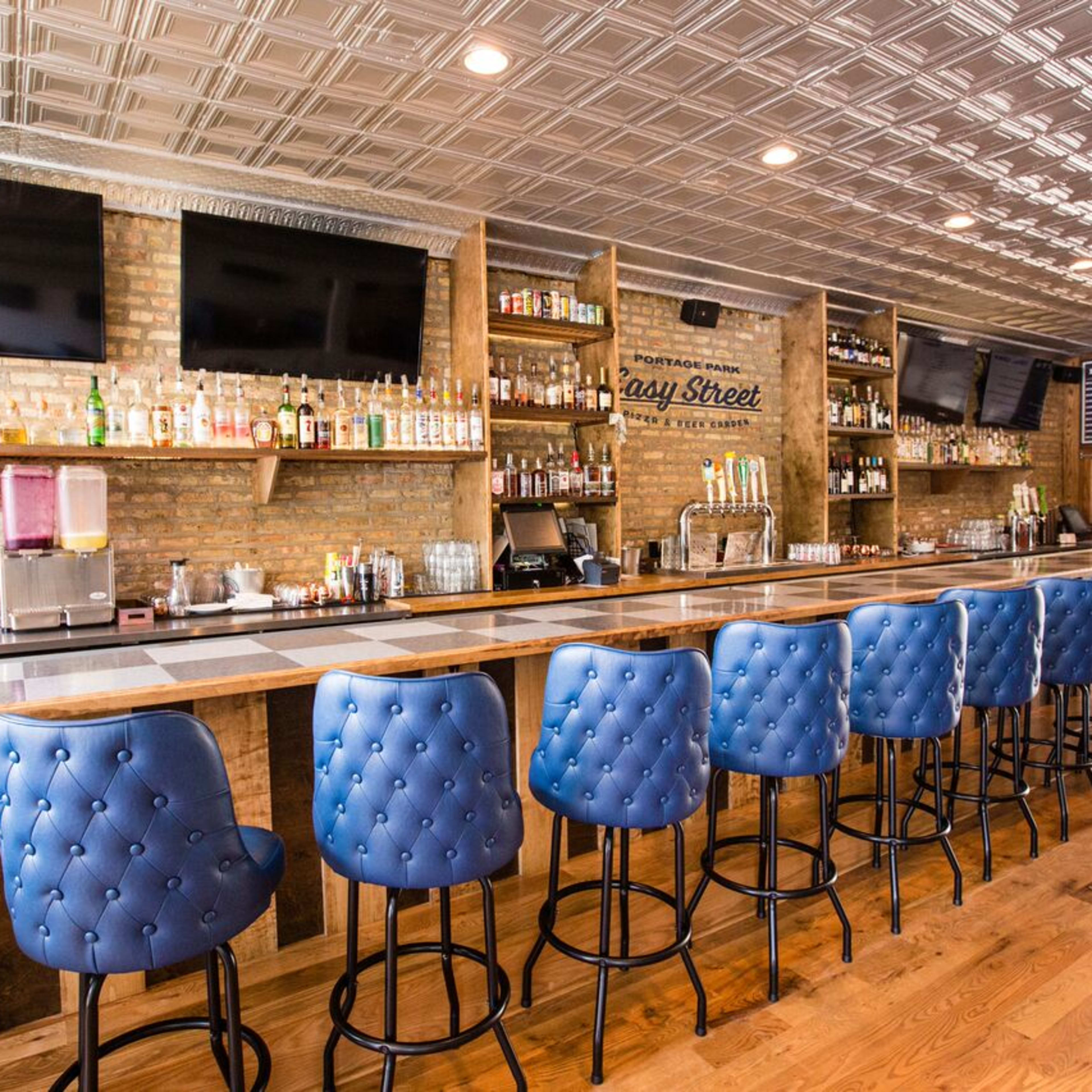 A modern bar with a long wooden counter, blue tufted bar stools, and various bottles displayed on shelves against a brick wall.