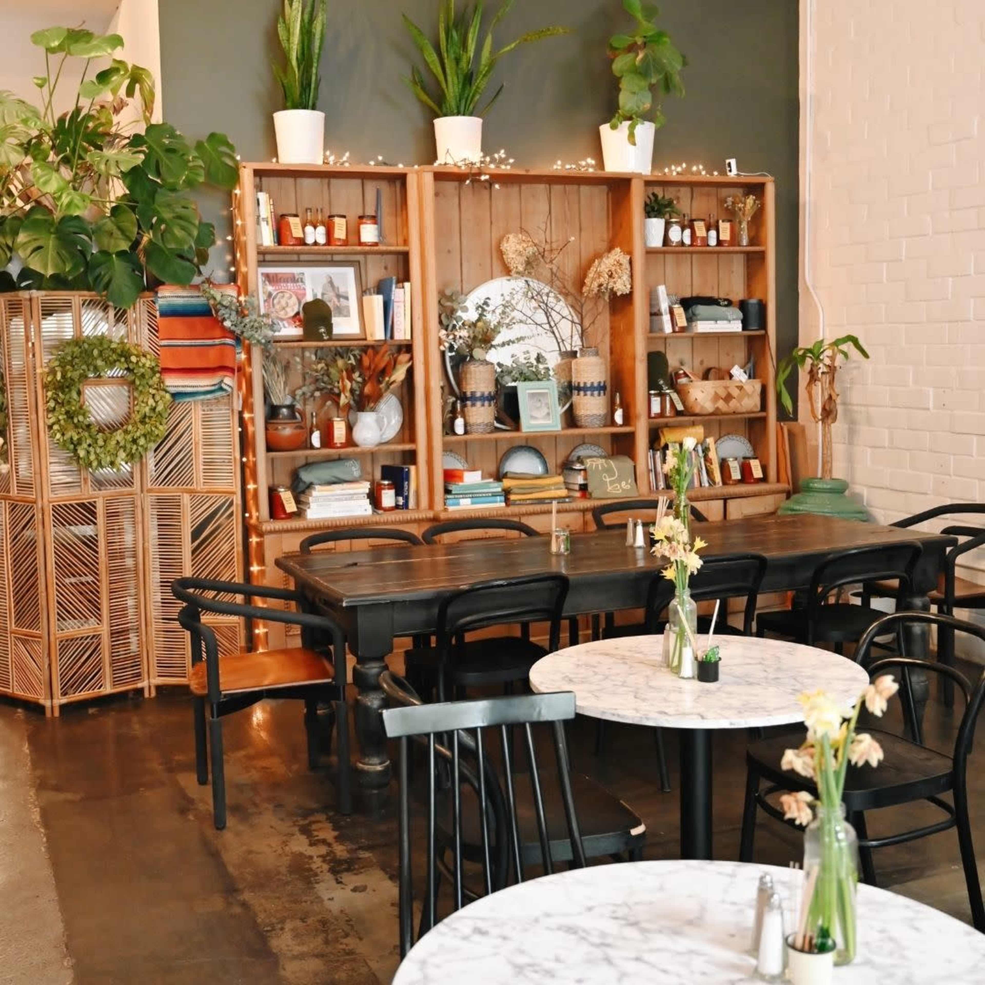 A cozy café interior featuring wooden and marble tables, shelving filled with decorative items, and greenery accenting the space.
