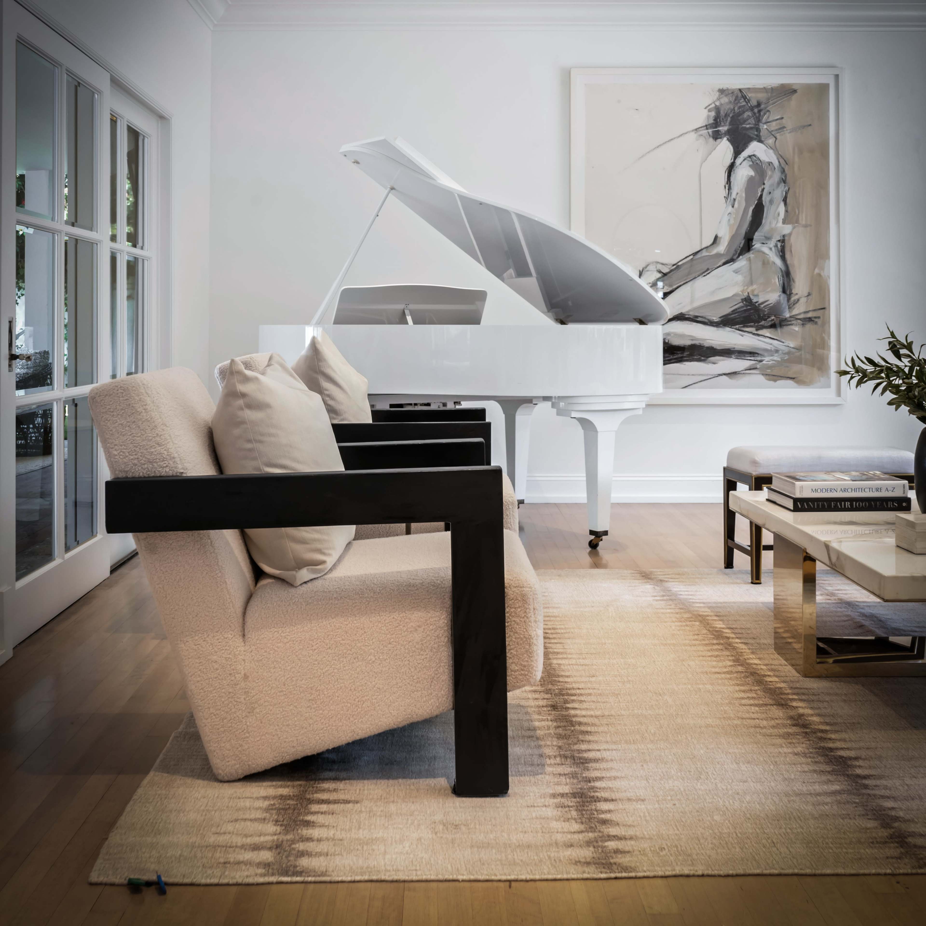 A white grand piano is positioned beside two modern armchairs in a brightly lit living room decorated with a large artwork and a coffee table.