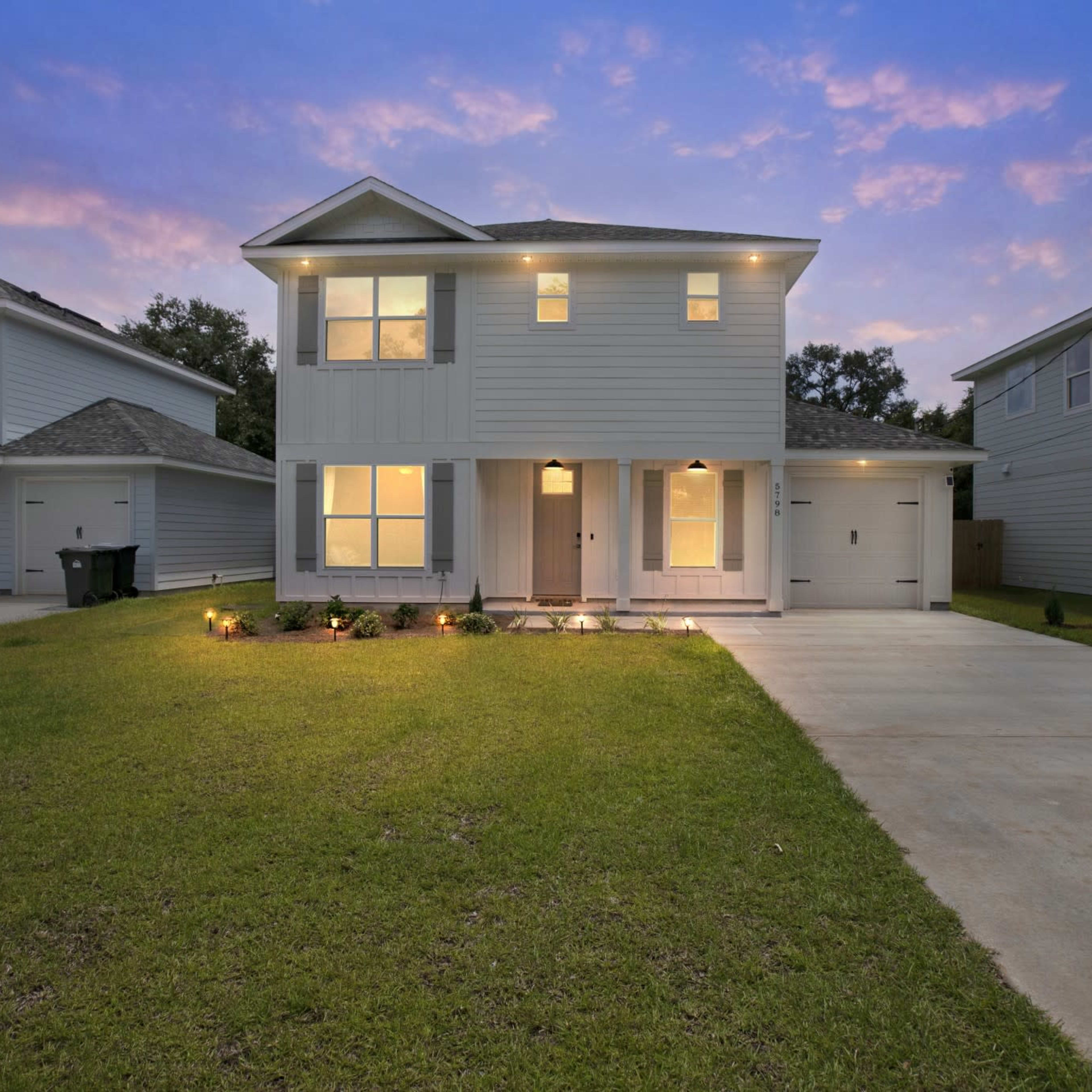 A two-story gray house with a covered front porch, large windows, and a concrete driveway is surrounded by a grassy yard at dusk.