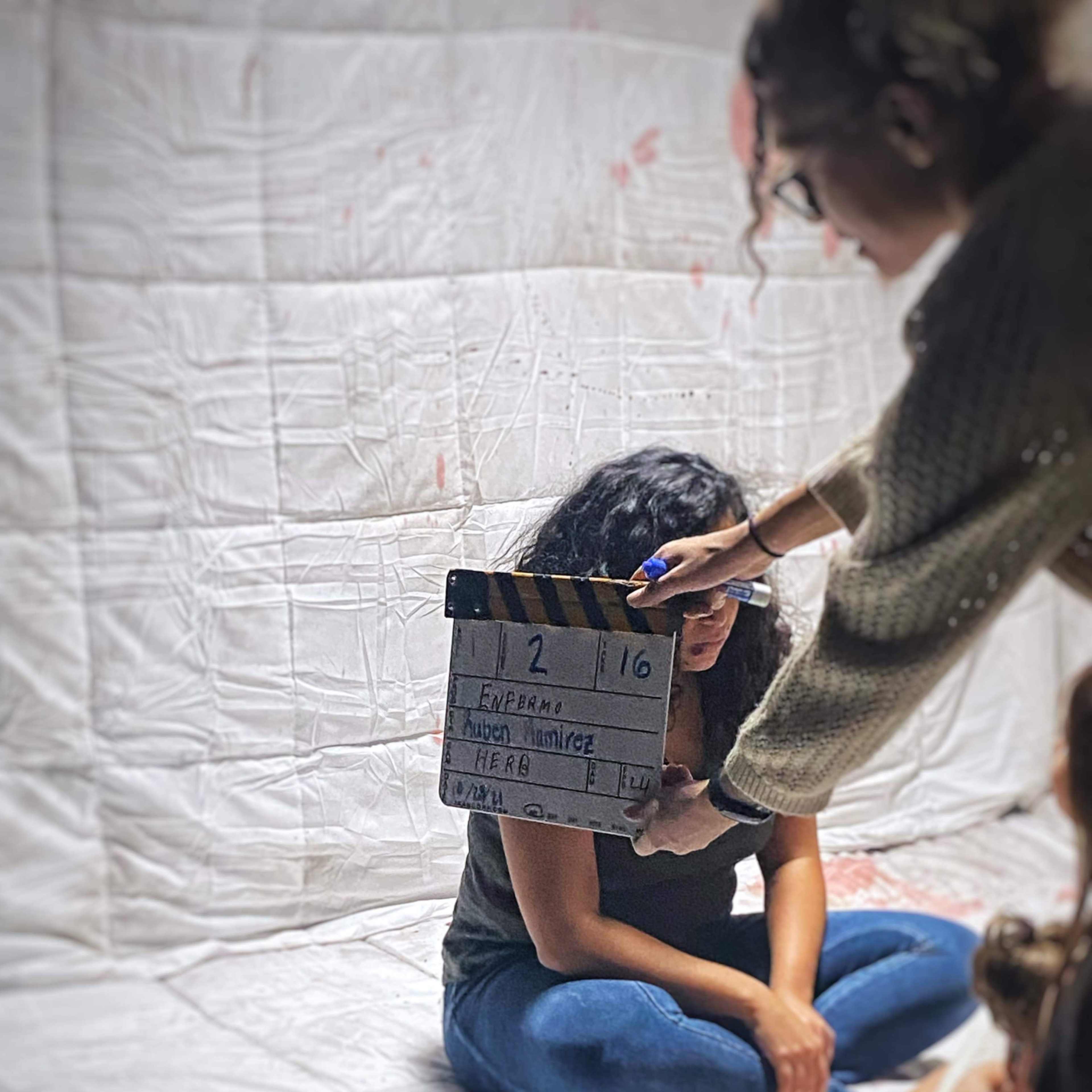 A woman sits on the floor against a white backdrop while another person holds a clapperboard labeled with details for a film scene.