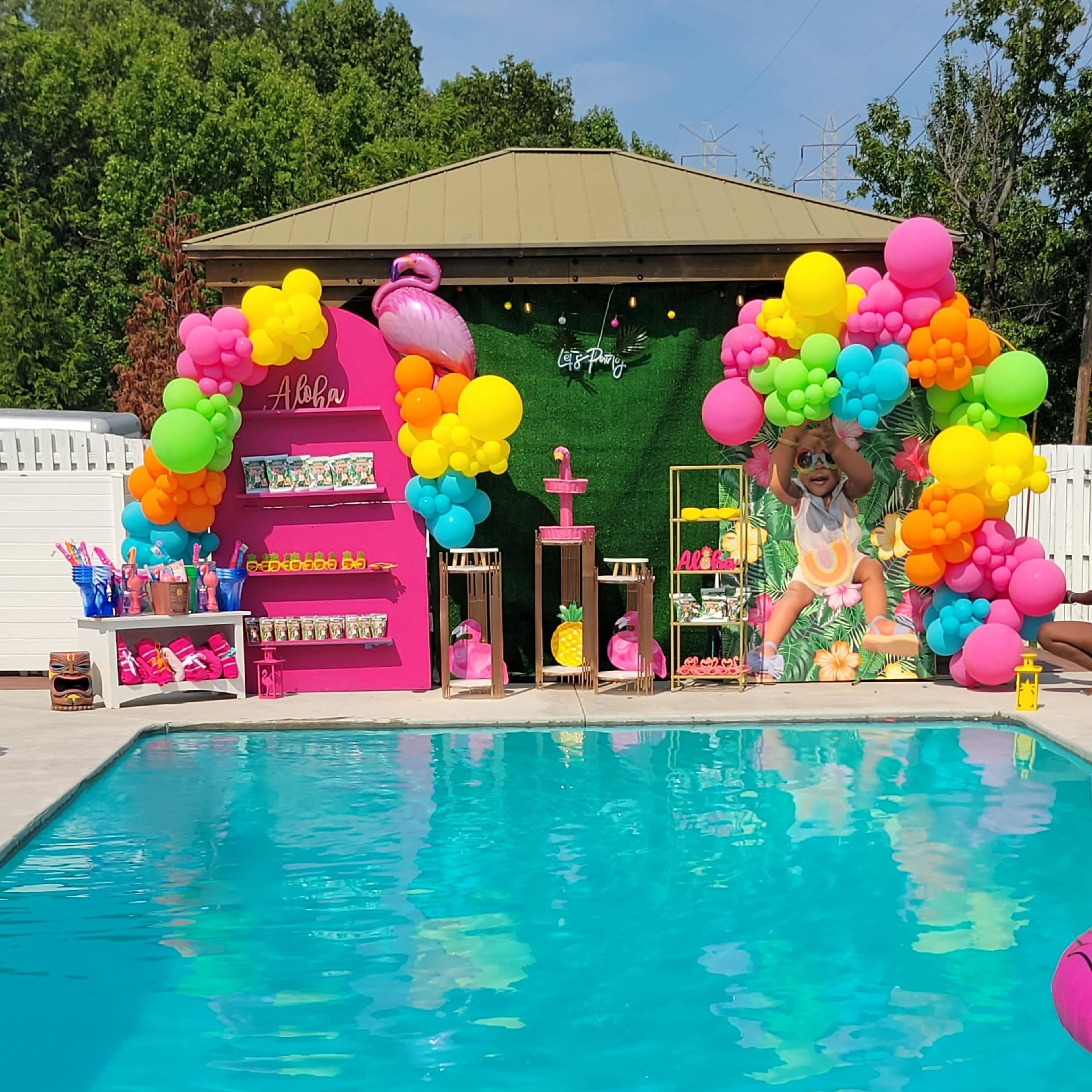 A vibrant pool area decorated with colorful balloons, a tropical-themed backdrop, and various inflatable toys surrounding the water.