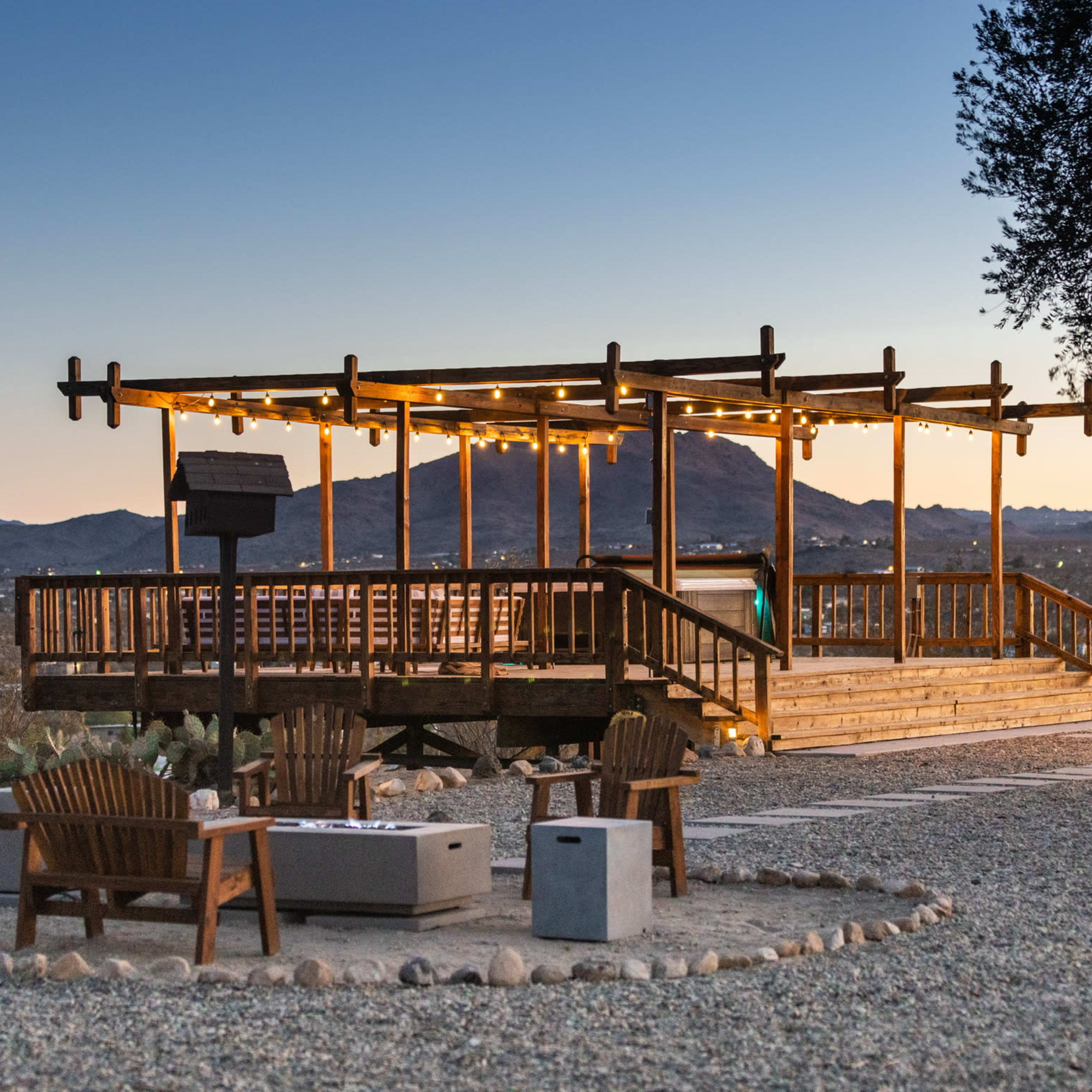 A wooden observation deck with string lights overlooks a valley surrounded by mountains, with stone fire pits and seating arranged at its base.
