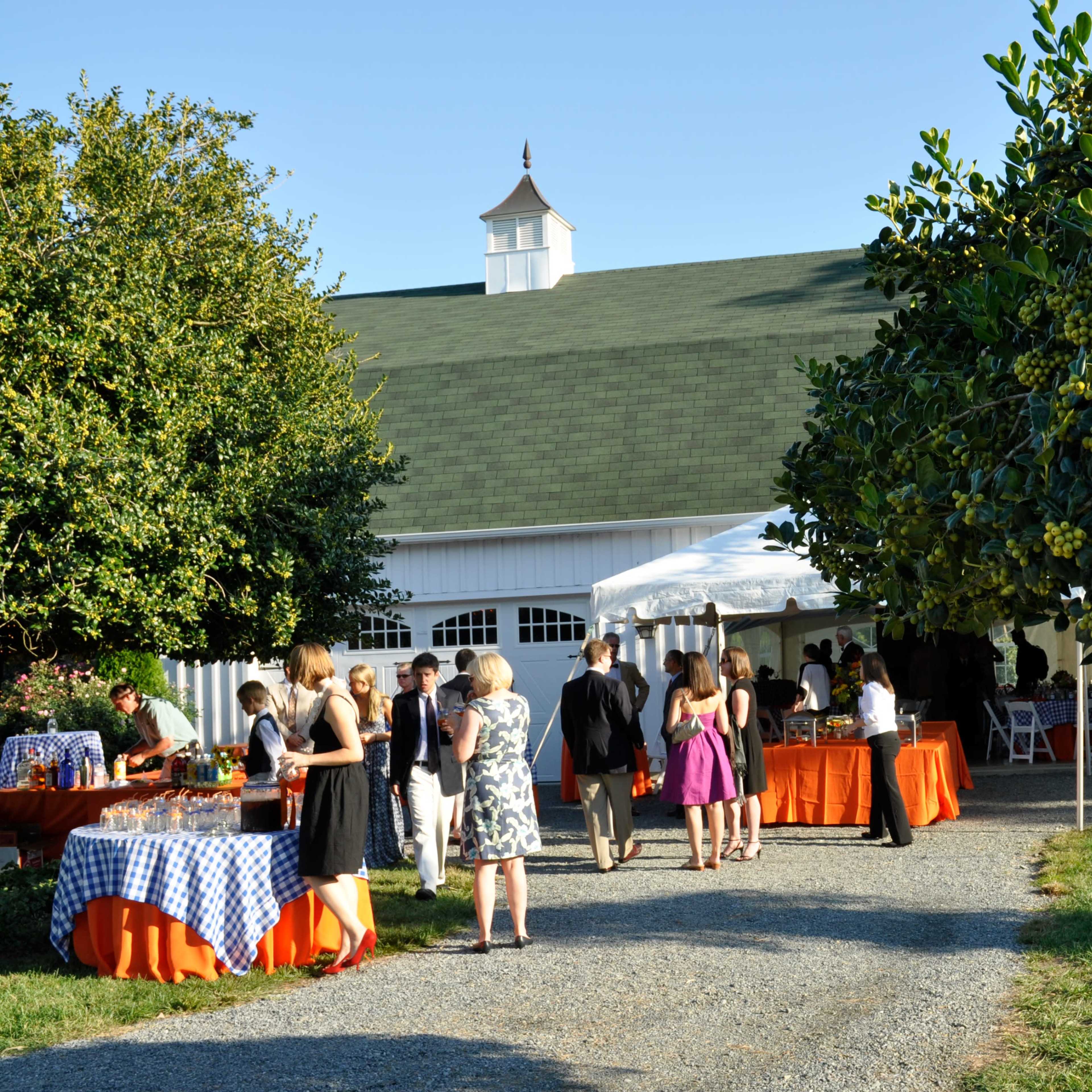 A group of people mingles outside a barn-style building during a social event, with tables set under a tent and a tree in the foreground.