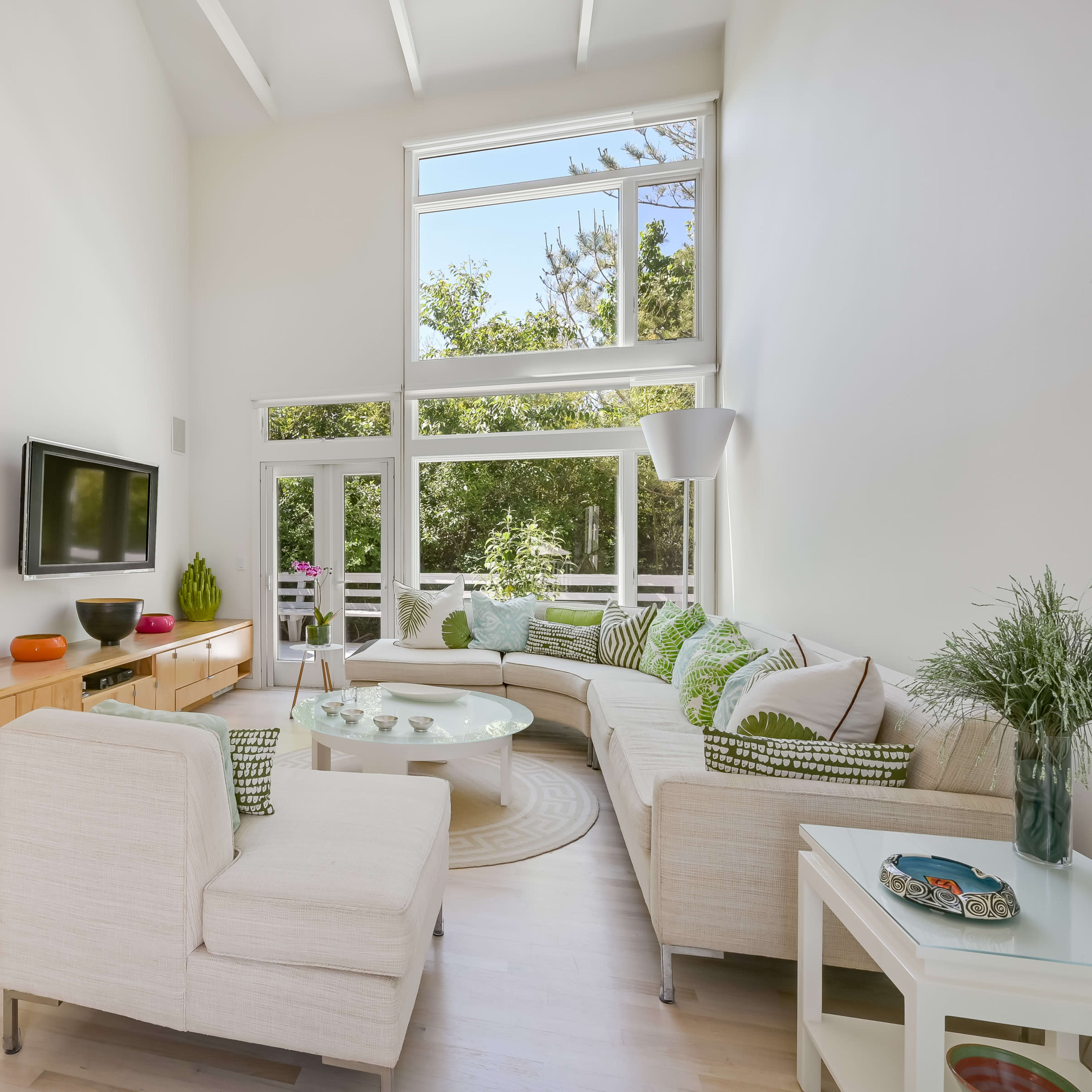 A bright living room features a sectional sofa, a television mounted on the wall, and large windows overlooking greenery.
