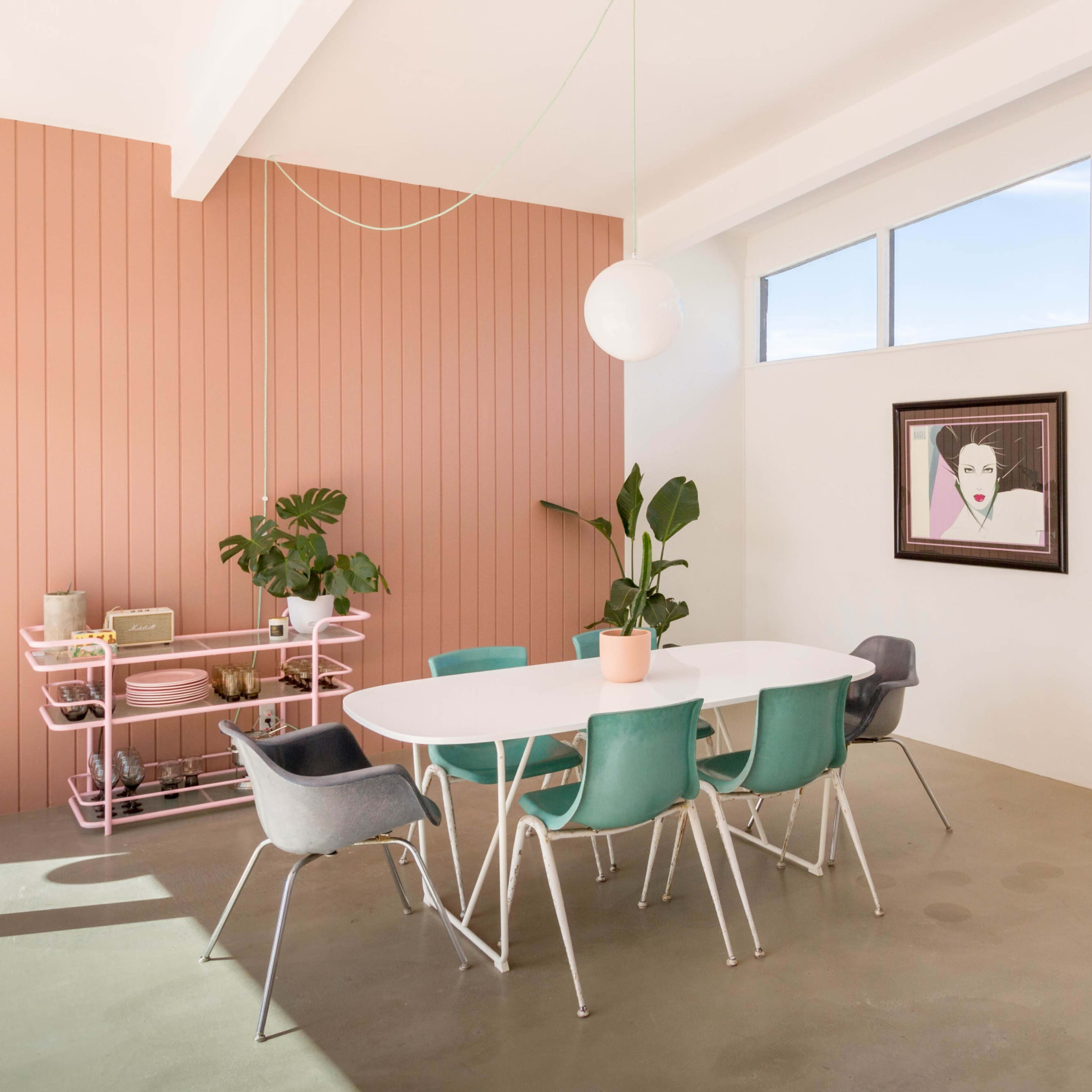 A modern dining area features a round white table surrounded by colorful chairs, with a pink wall, a large window, and a mirror reflecting a poolside view.