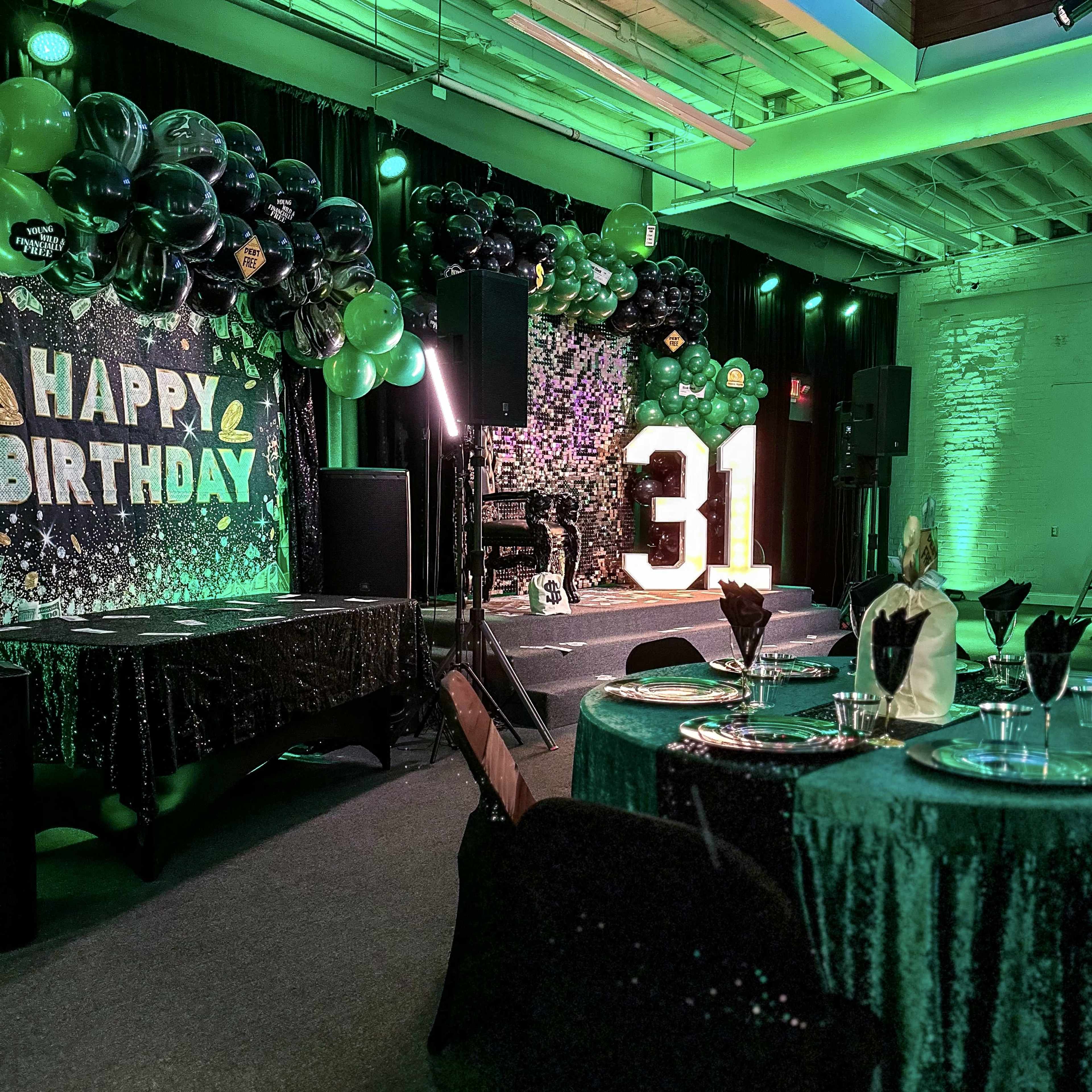 A birthday party setup featuring a "Happy Birthday" backdrop, green and black decorations, and a large illuminated "31" beside elegantly arranged tables with black and green table settings.