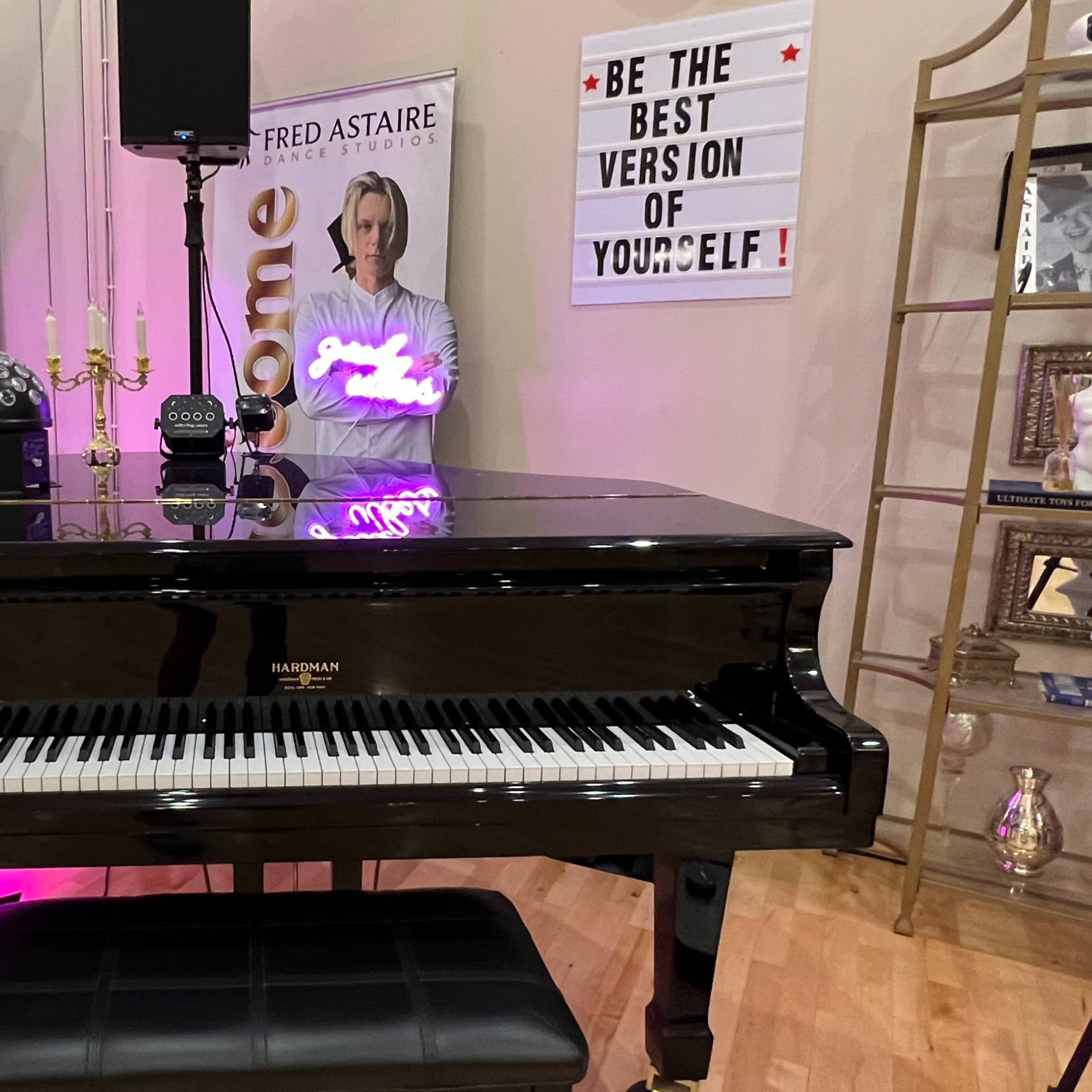 A black grand piano in a studio setting, accompanied by mirrors, decorative shelves, and a motivational sign on the wall.