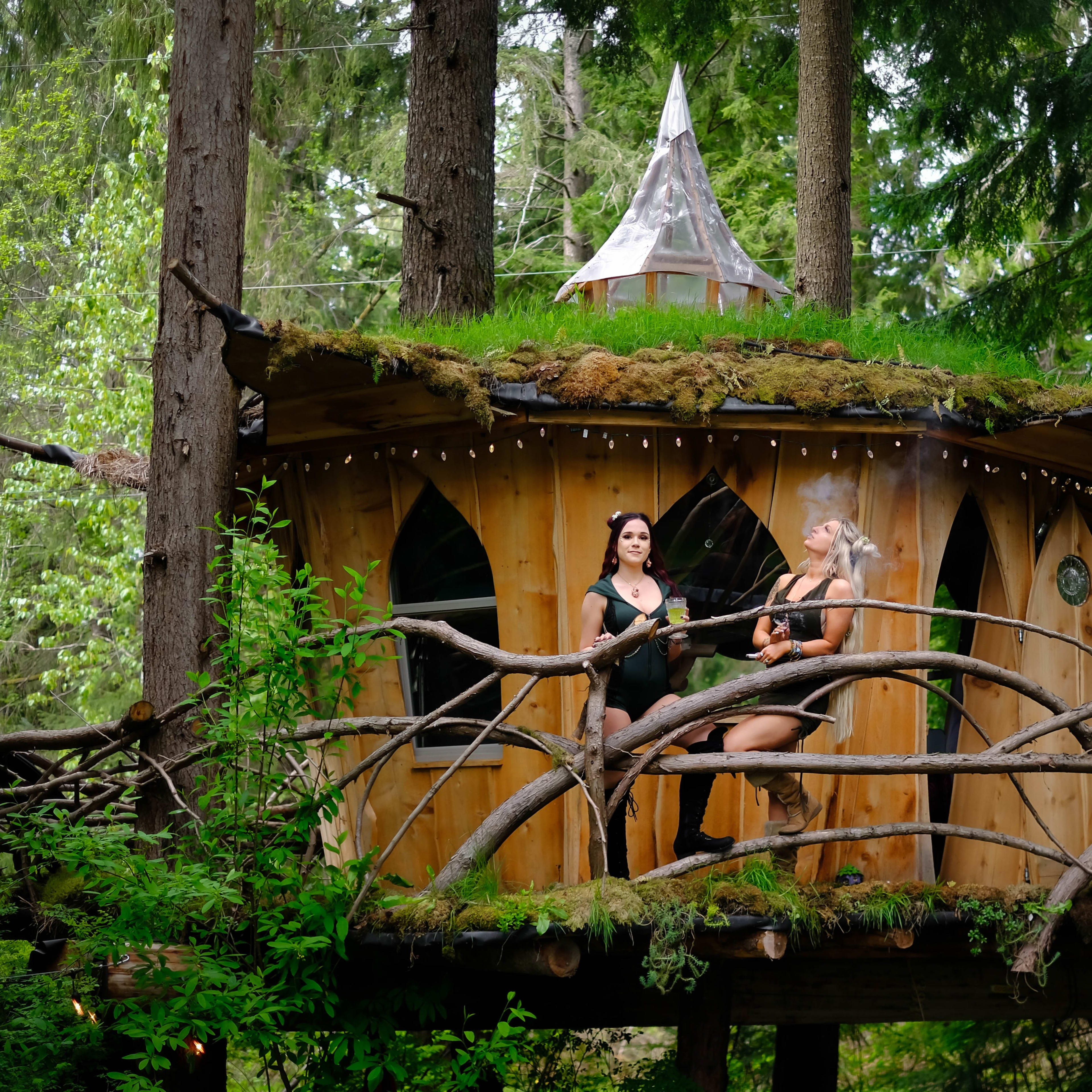 Two women are playing instruments on the balcony of a wooden treehouse surrounded by dense greenery.