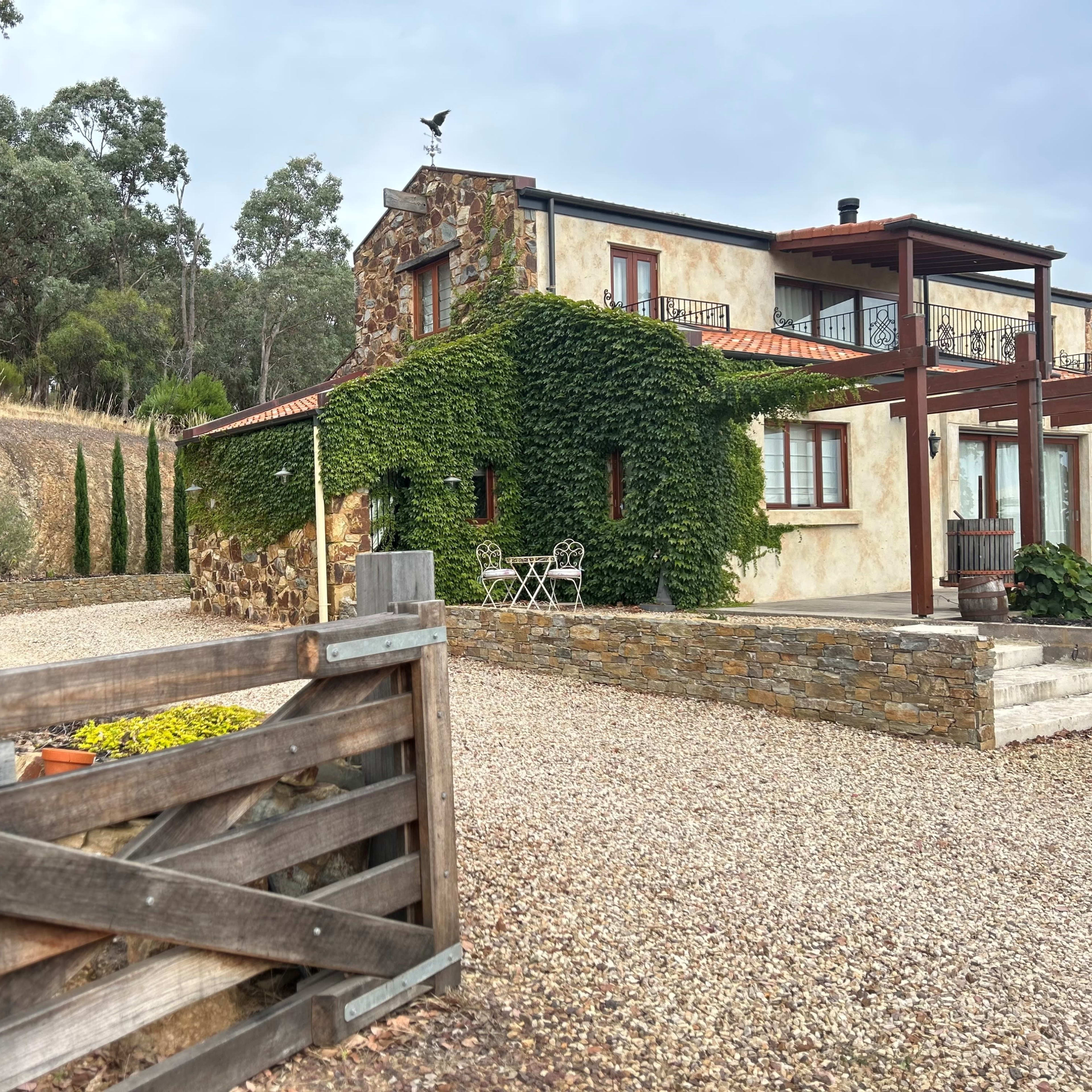 A stone and stucco house covered in greenery sits at the end of a gravel driveway, surrounded by tall cypress trees and rolling hills.