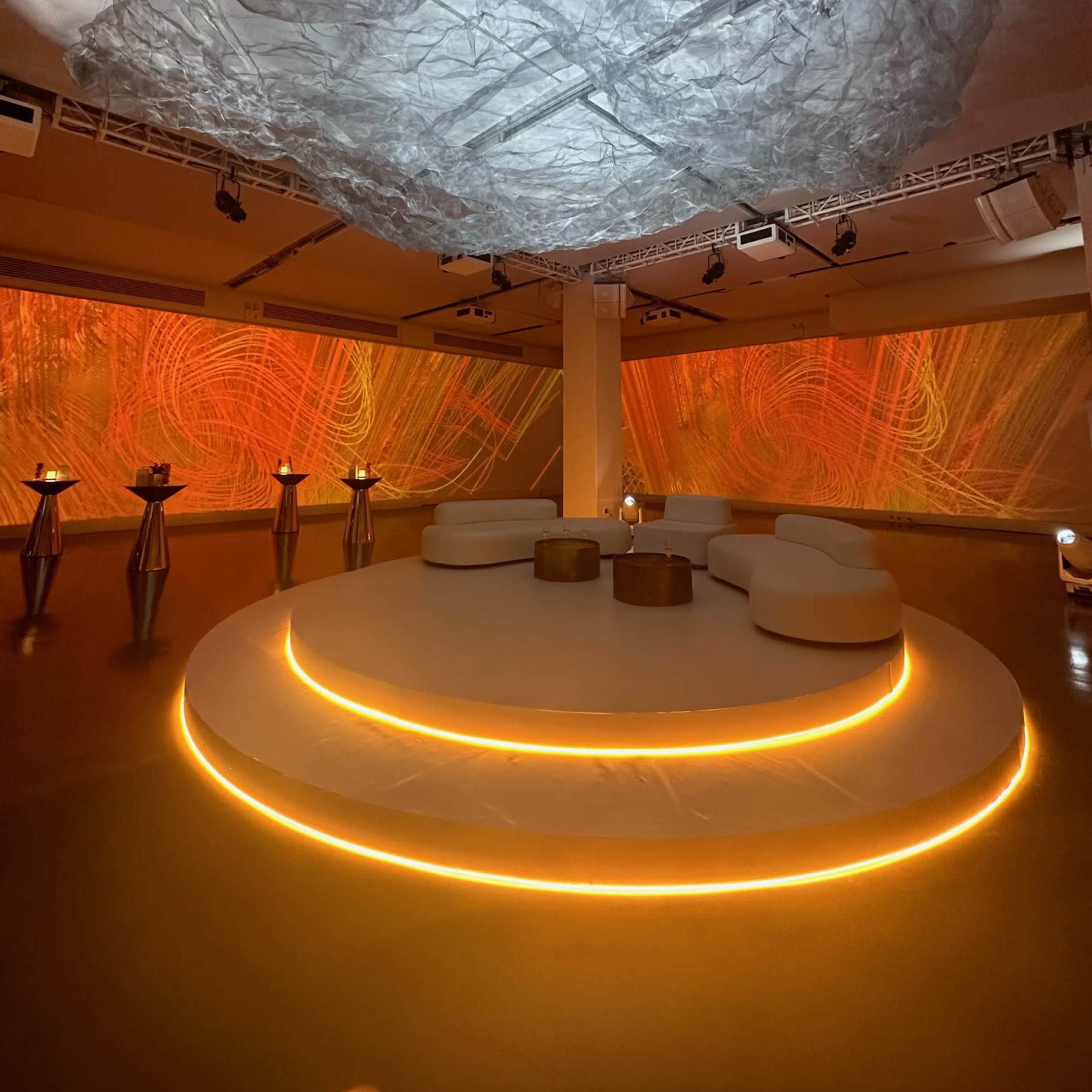 A modern interior space featuring a circular seating arrangement illuminated by orange lights, with abstract projections on the walls and a textured ceiling installation.