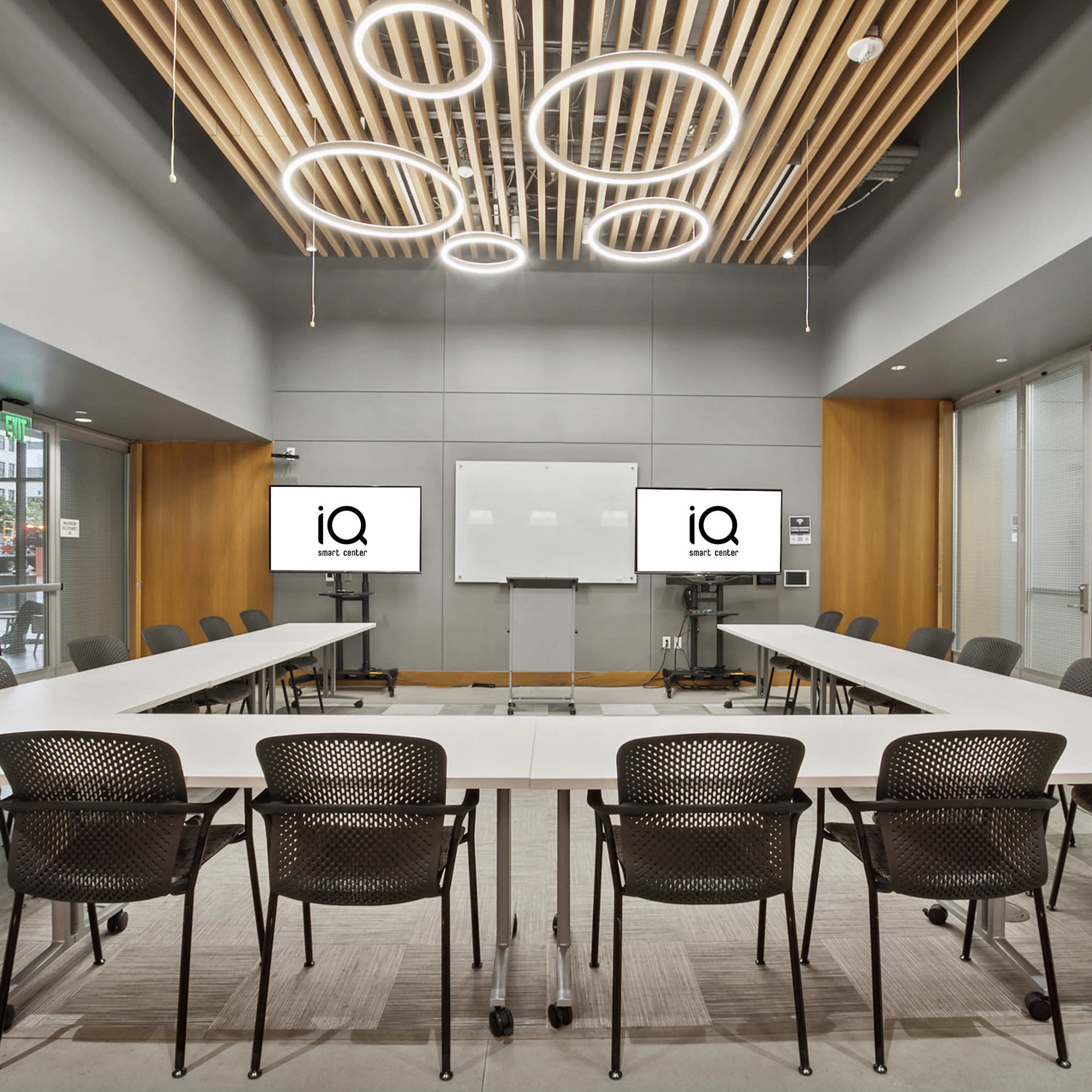 A modern conference room features a large oval table surrounded by black chairs, with two screens displaying the logo "iQ" and a whiteboard on the wall.