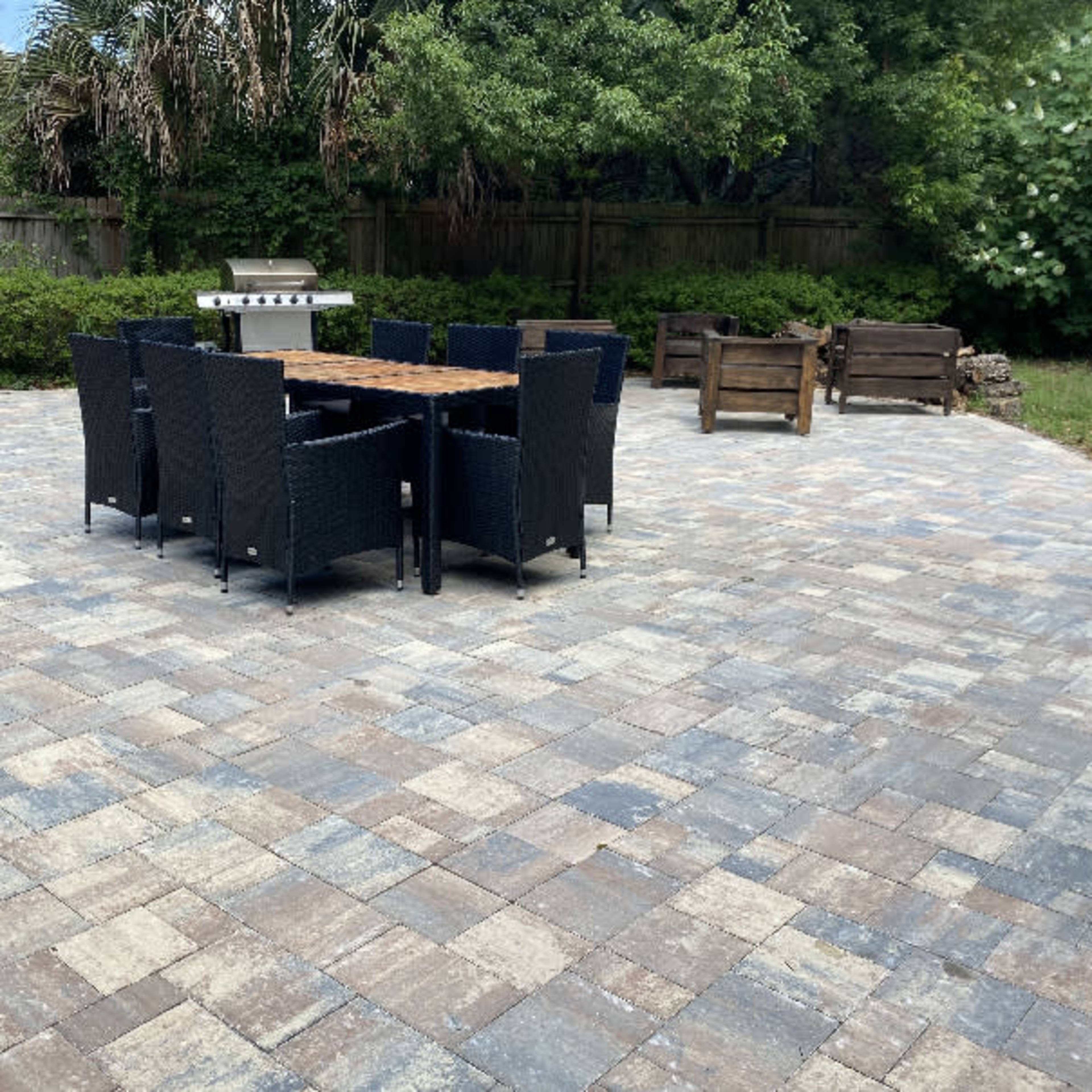 A patio area features a rectangular table surrounded by black wicker chairs, with a grill in the background and a landscaped border of shrubs and trees.