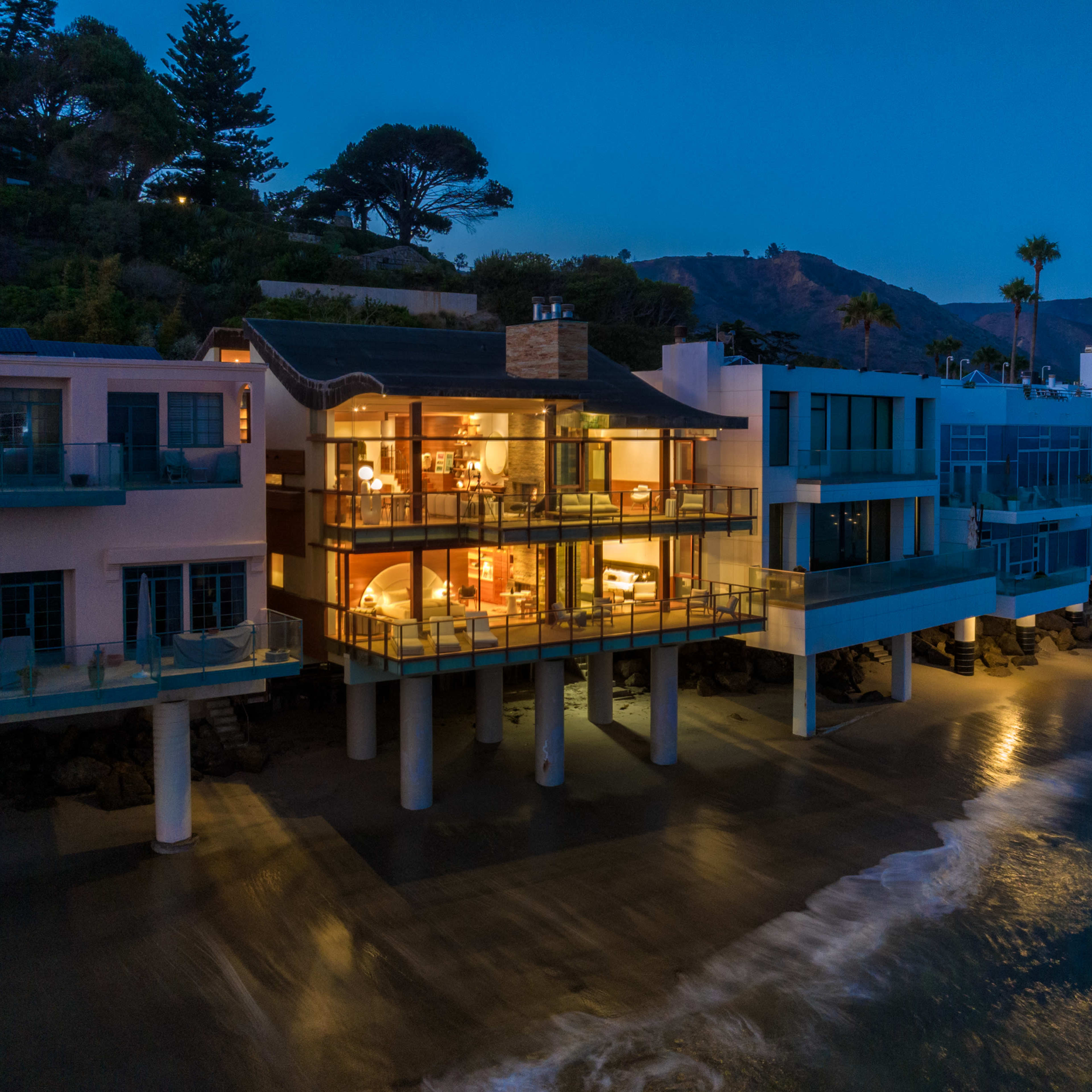 A modern beachfront house with large glass windows is illuminated at night, set against a mountainous backdrop by the sea.