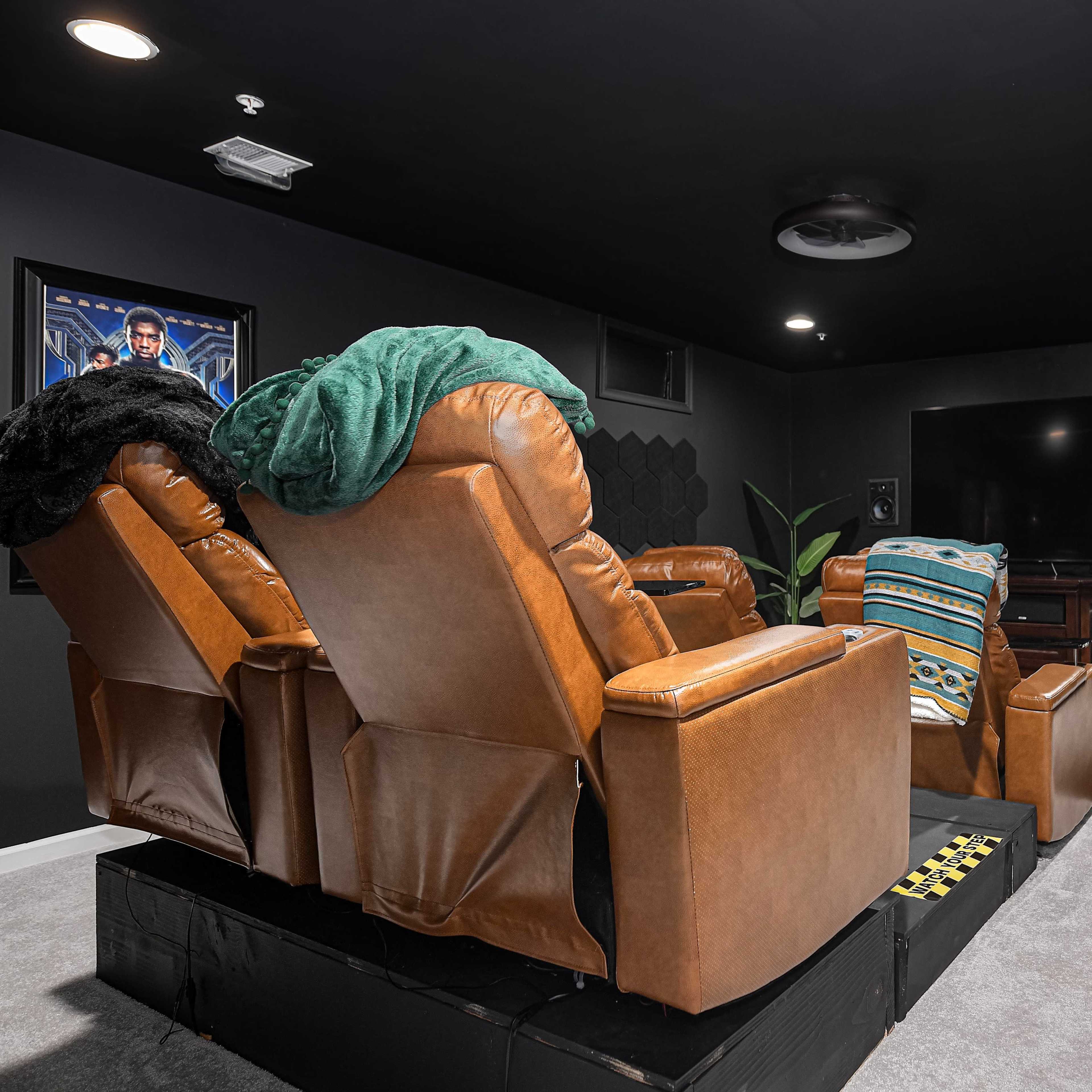 A home theater room with two brown reclining chairs facing a large wall-mounted television, surrounded by dark walls and minimal decor.