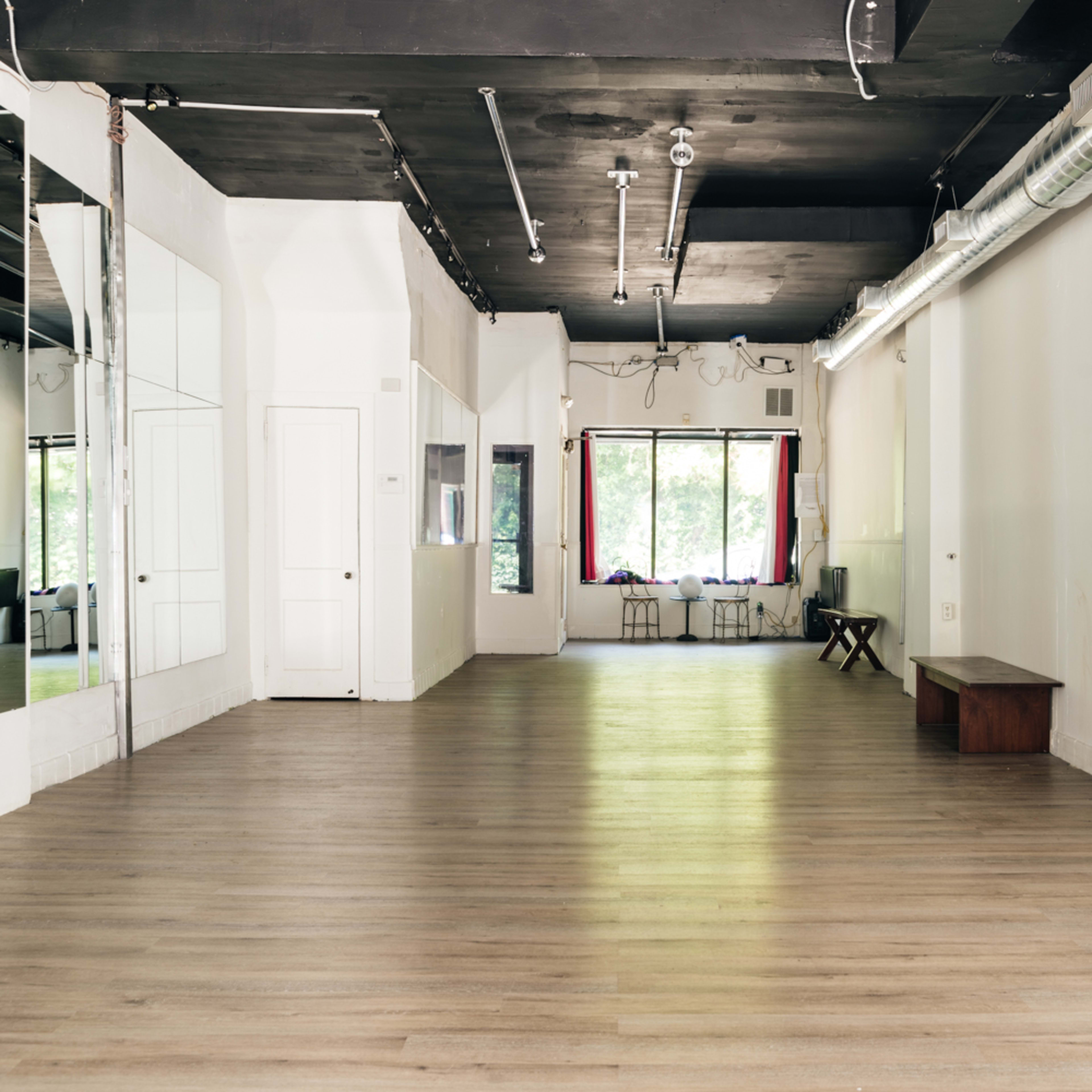 An empty dance studio with hardwood floors, large mirrors on one wall, and natural light streaming through a window at the far end.