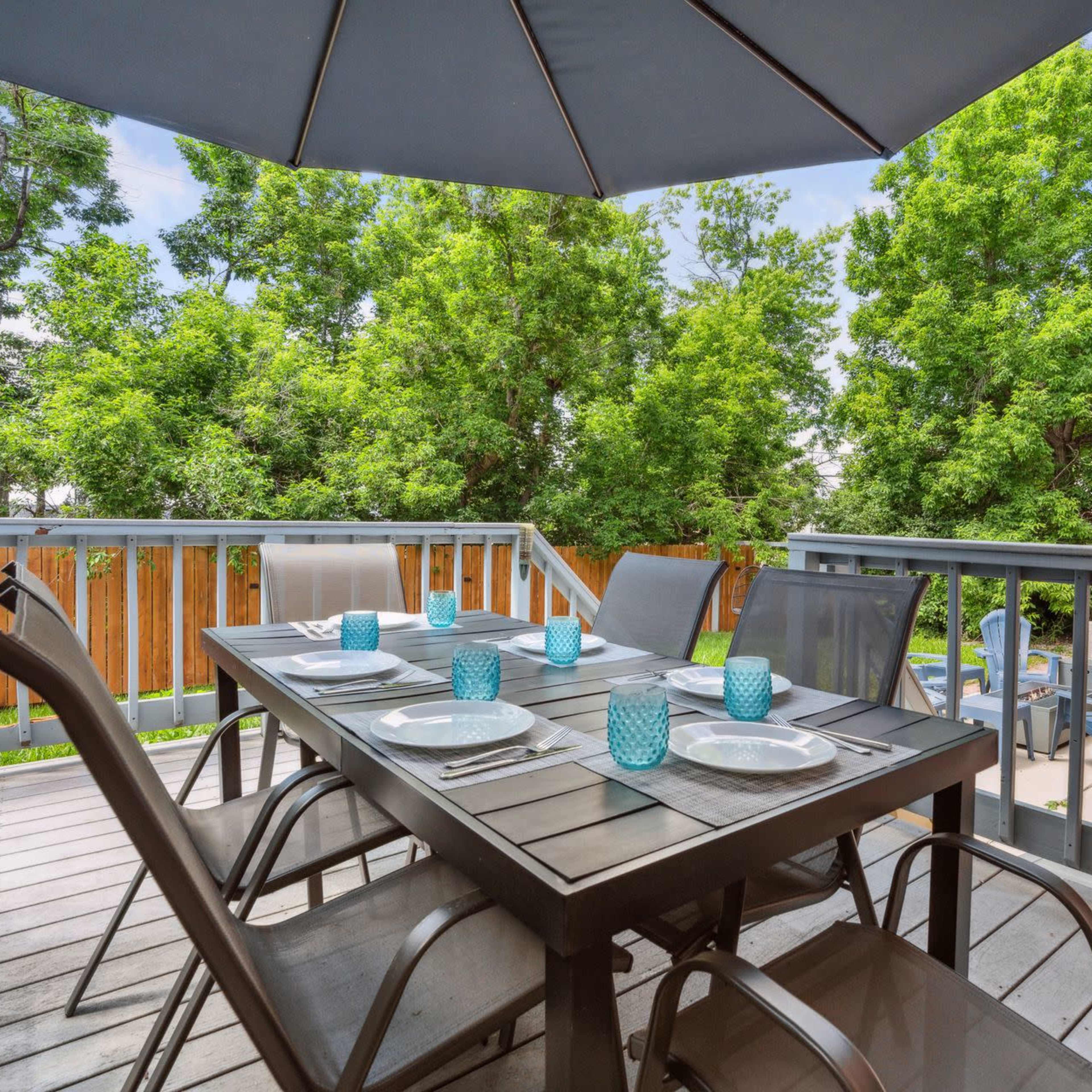 A dining table with four chairs is set on a wooden deck under a gray umbrella, surrounded by greenery and a fenced yard.