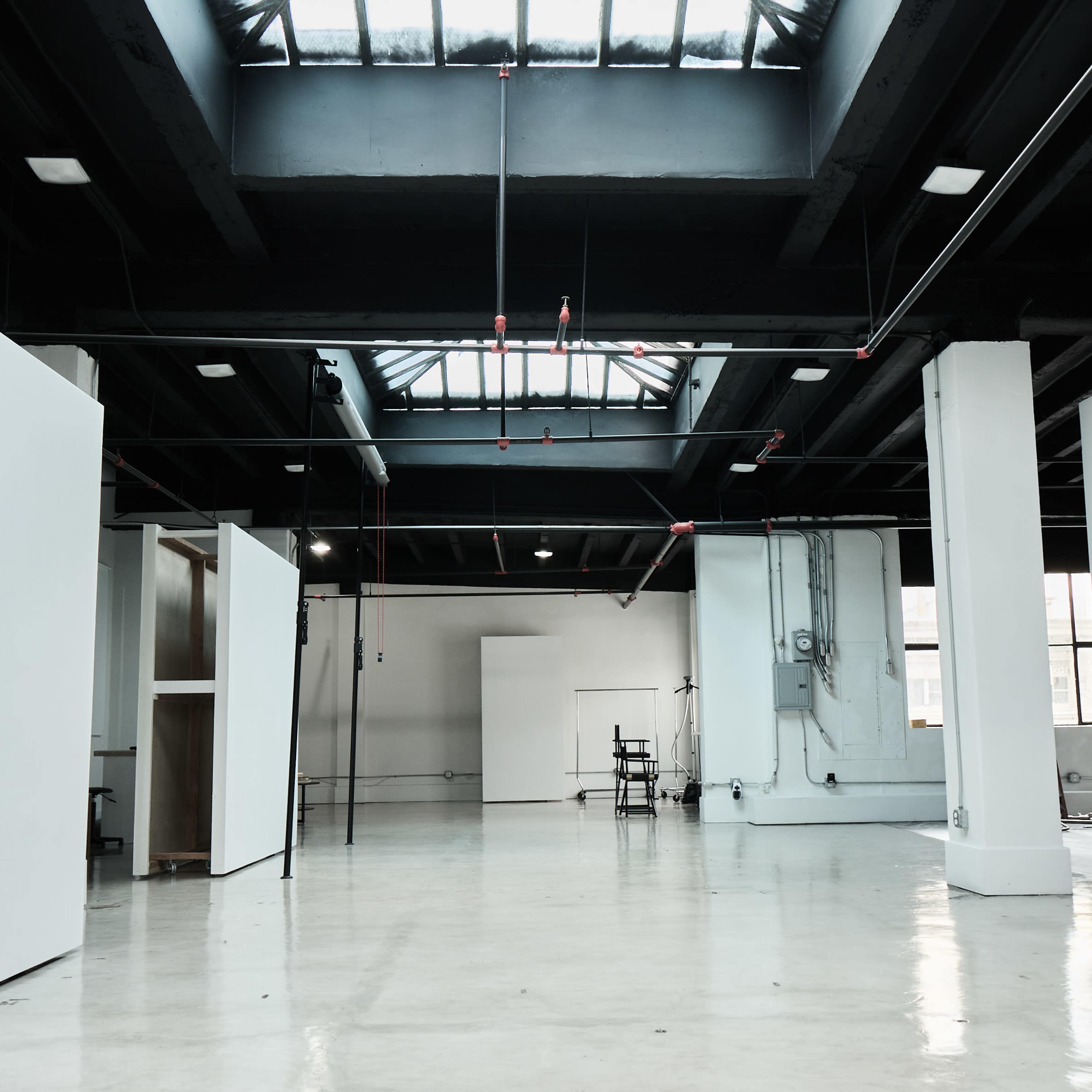 A spacious, industrial-style room with large windows, white walls, and a polished concrete floor, featuring minimal furnishings and scattered lighting.