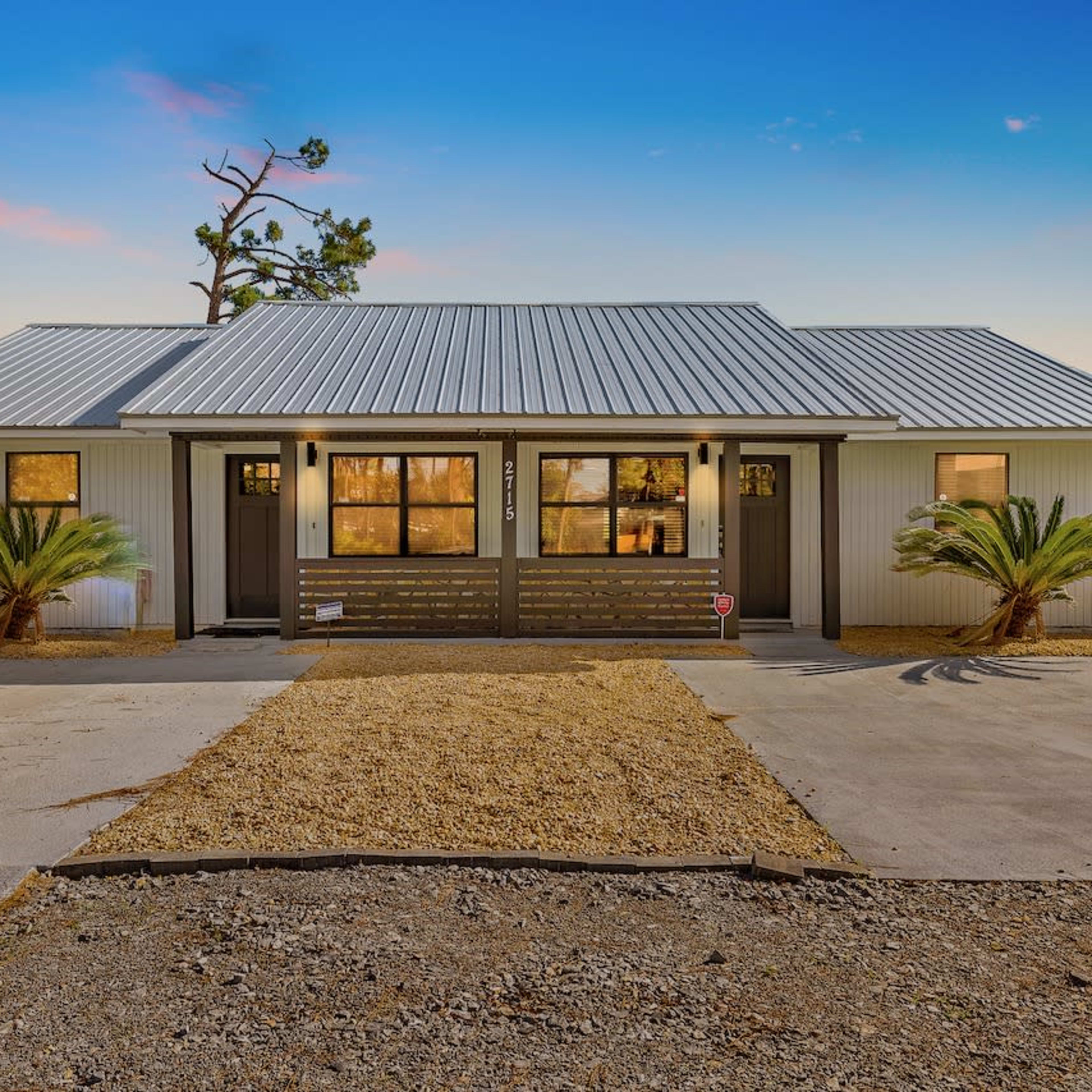 A modern, single-story duplex with a metal roof, palm trees in the front yard, and a gravel driveway.