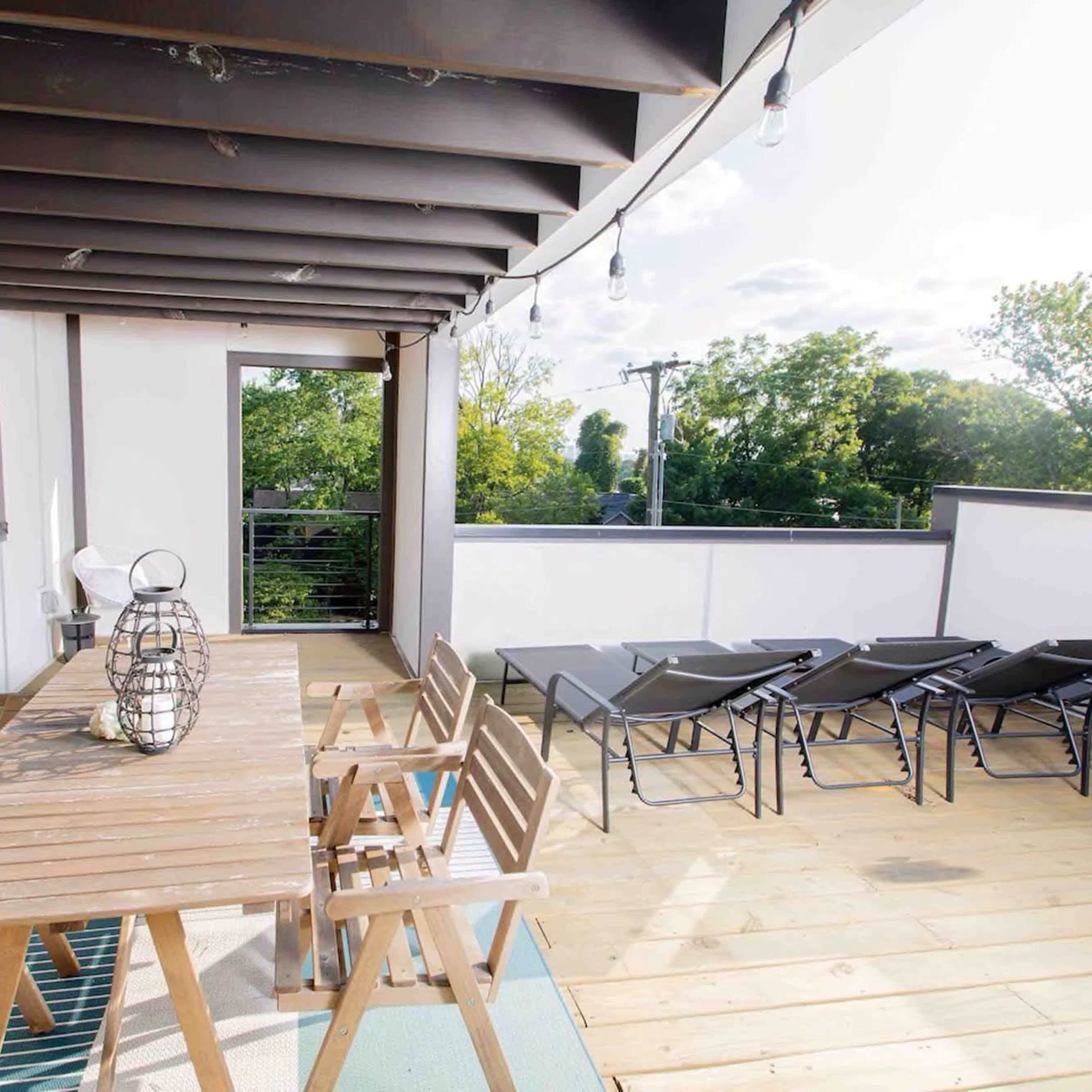 A wooden dining table surrounded by chairs is situated on a terrace with lounge chairs and greenery visible in the background.