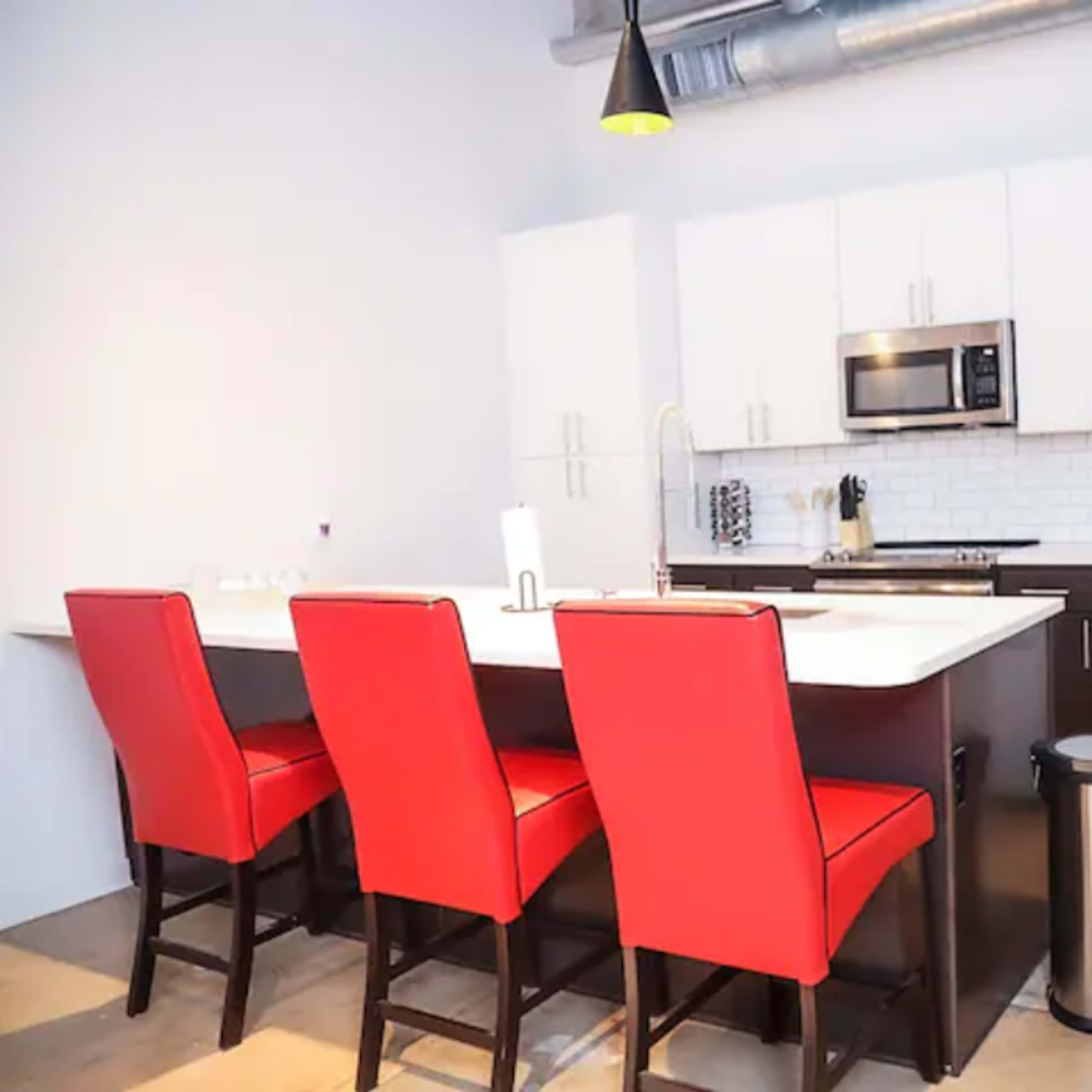 A modern kitchen with white cabinets, a stainless steel refrigerator, a microwave, and four red bar stools at a central island.