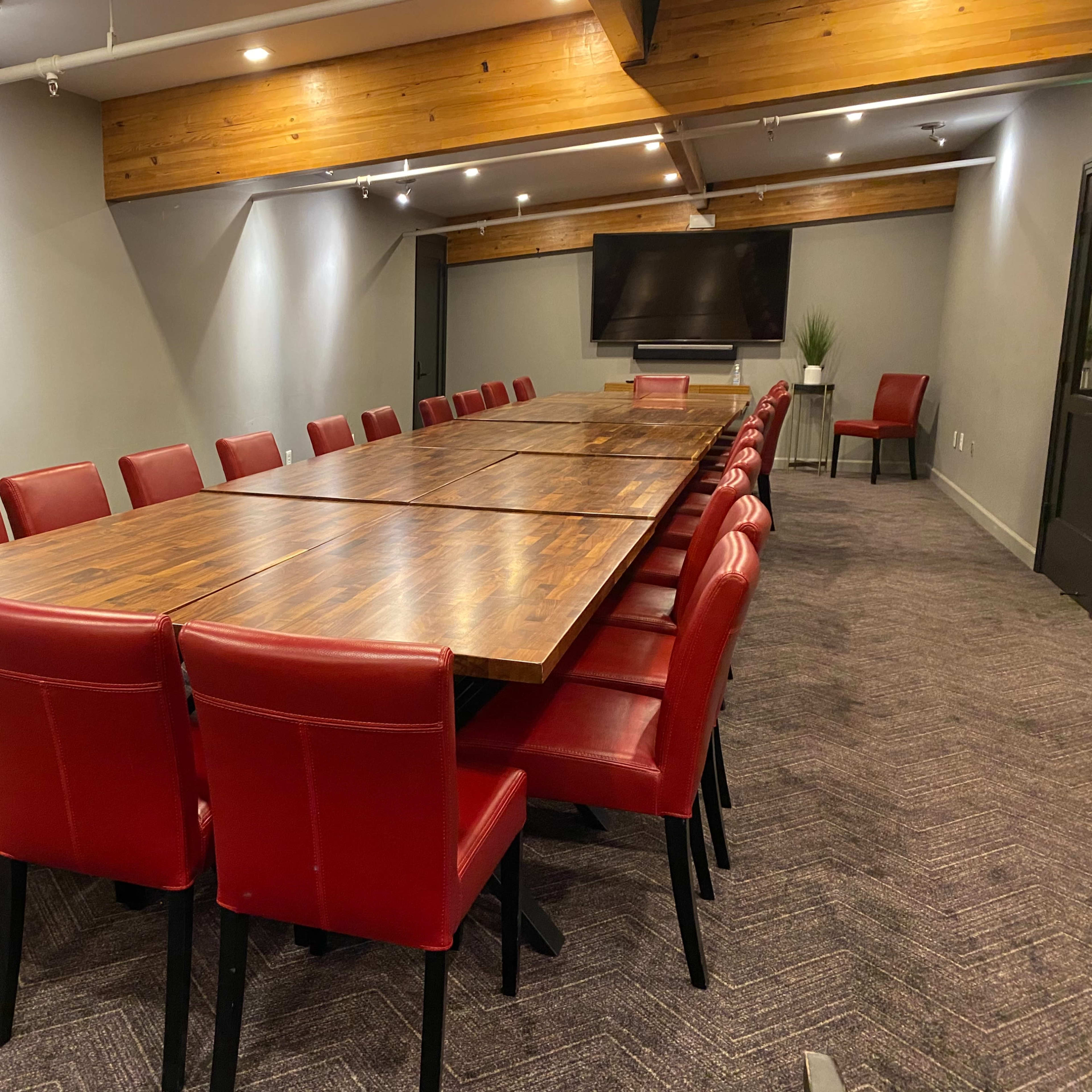 A long conference table surrounded by red chairs is situated in a meeting room with gray walls and wooden accents.