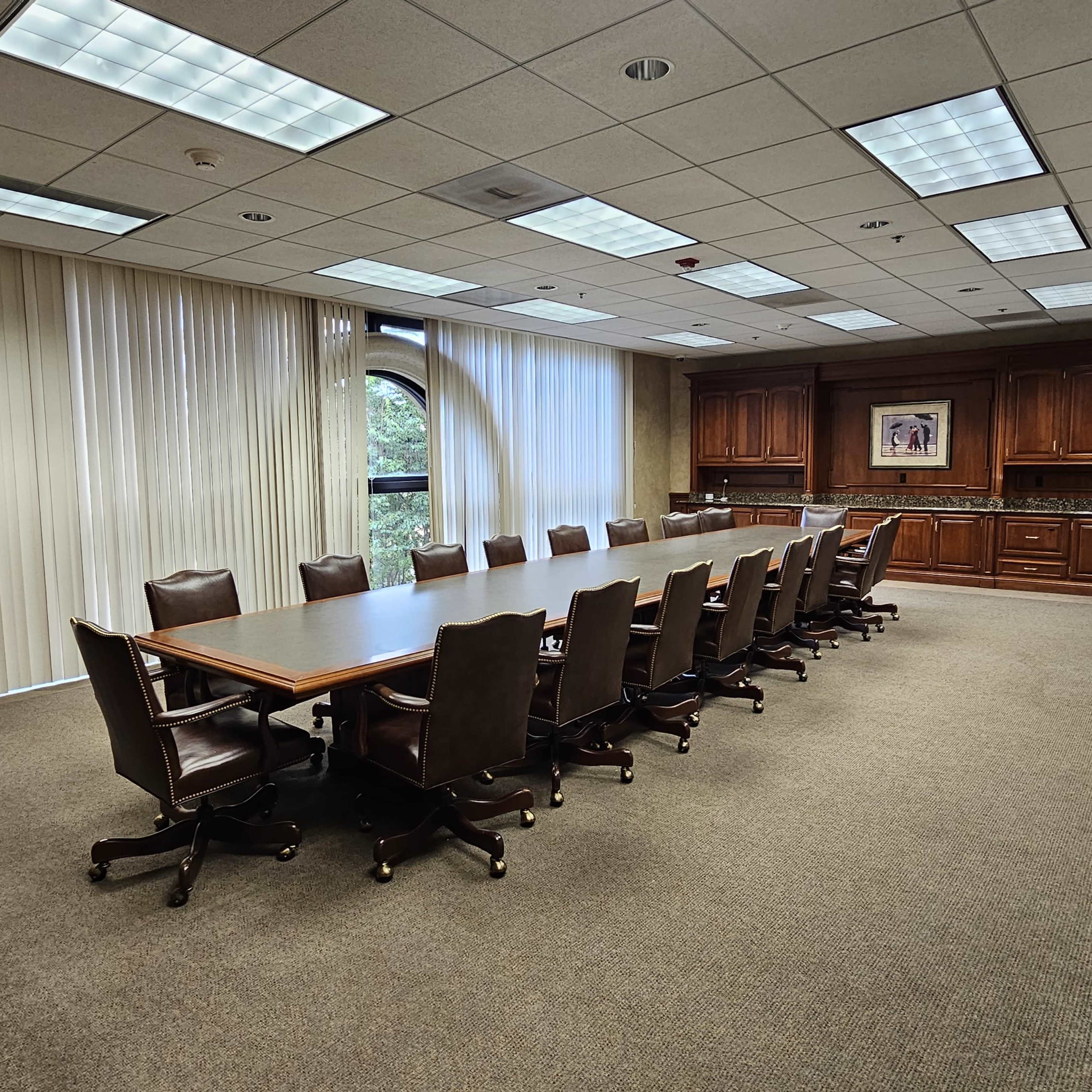 A large conference room with a long wooden table surrounded by leather chairs and a kitchenette area in the background.