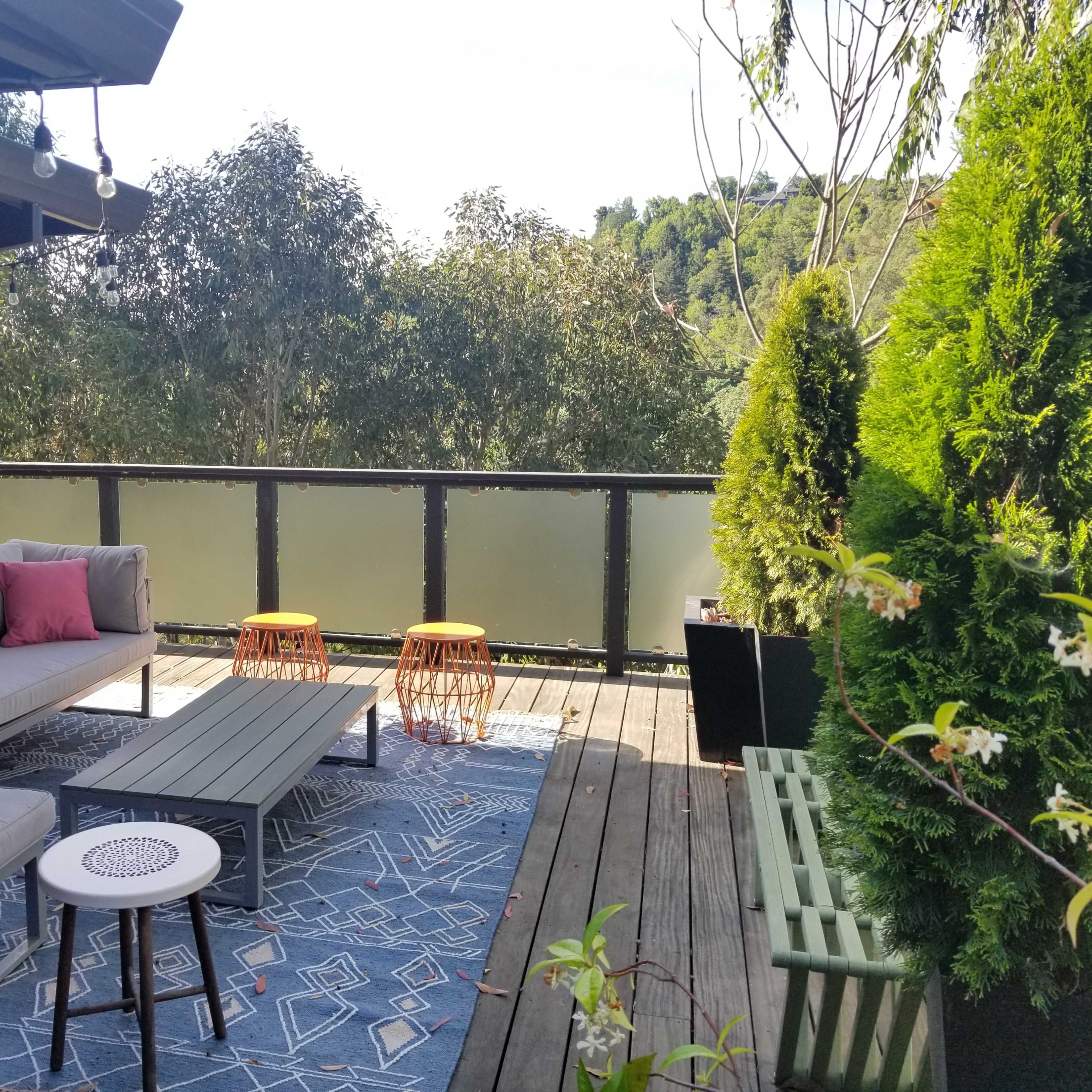 The scene features a terrace with a gray couch, two orange stools, and a coffee table, surrounded by greenery and a railing with a view of trees in the background.