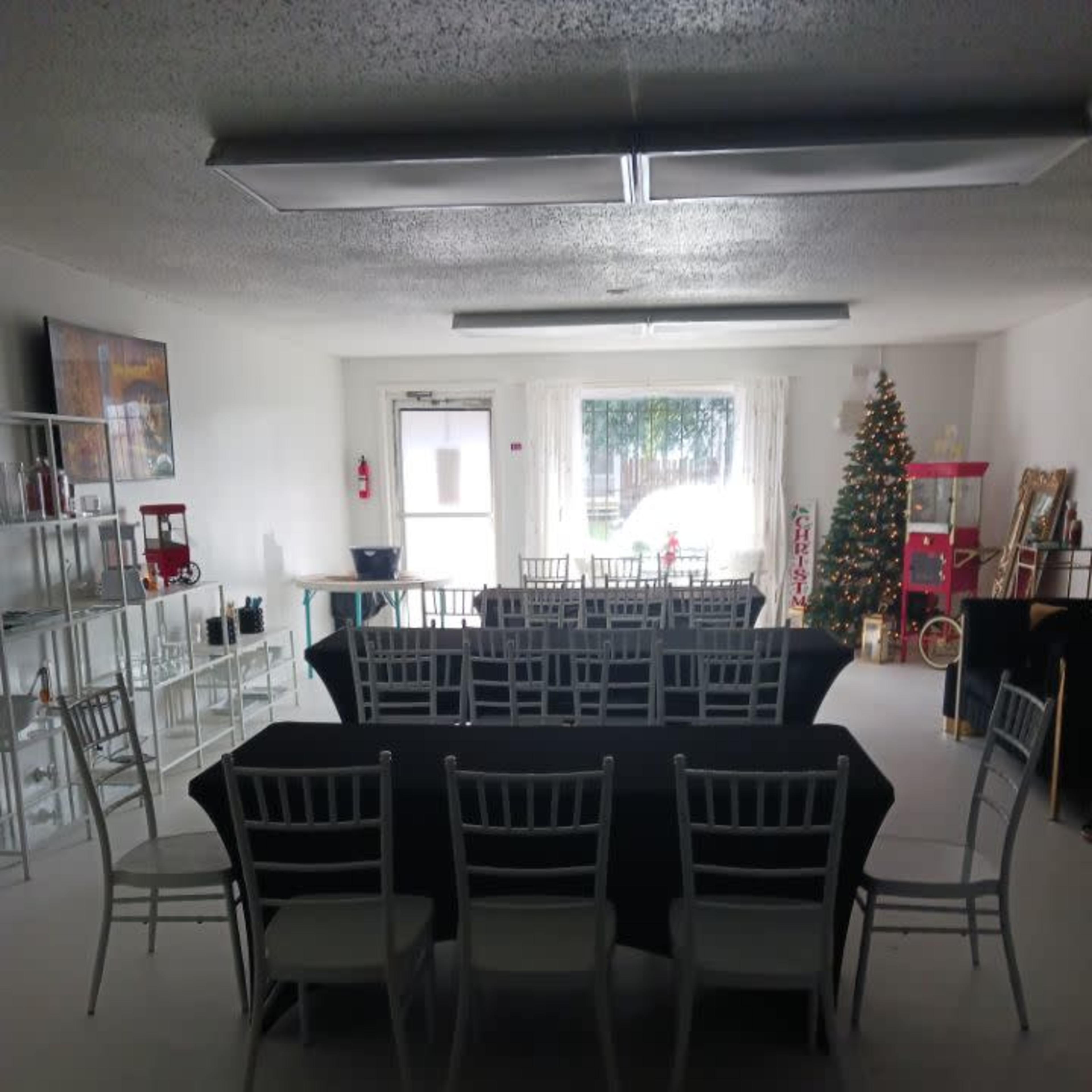 A spacious room set up for an event, featuring multiple tables covered with black cloth and surrounded by white chairs, with a decorated Christmas tree in one corner.