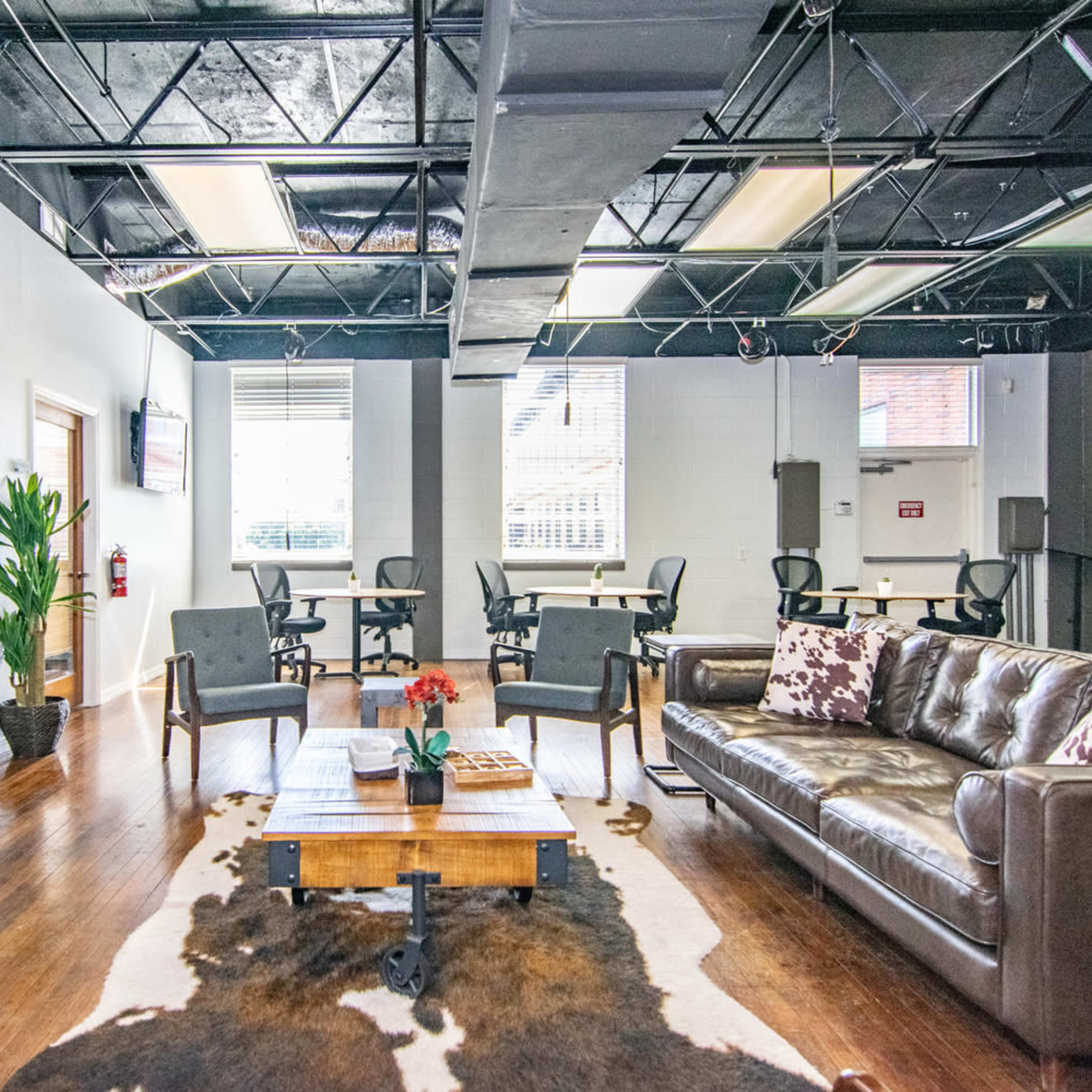 A modern office lounge features a leather sofa, a wooden coffee table, and a large television, with desks set up in the background.