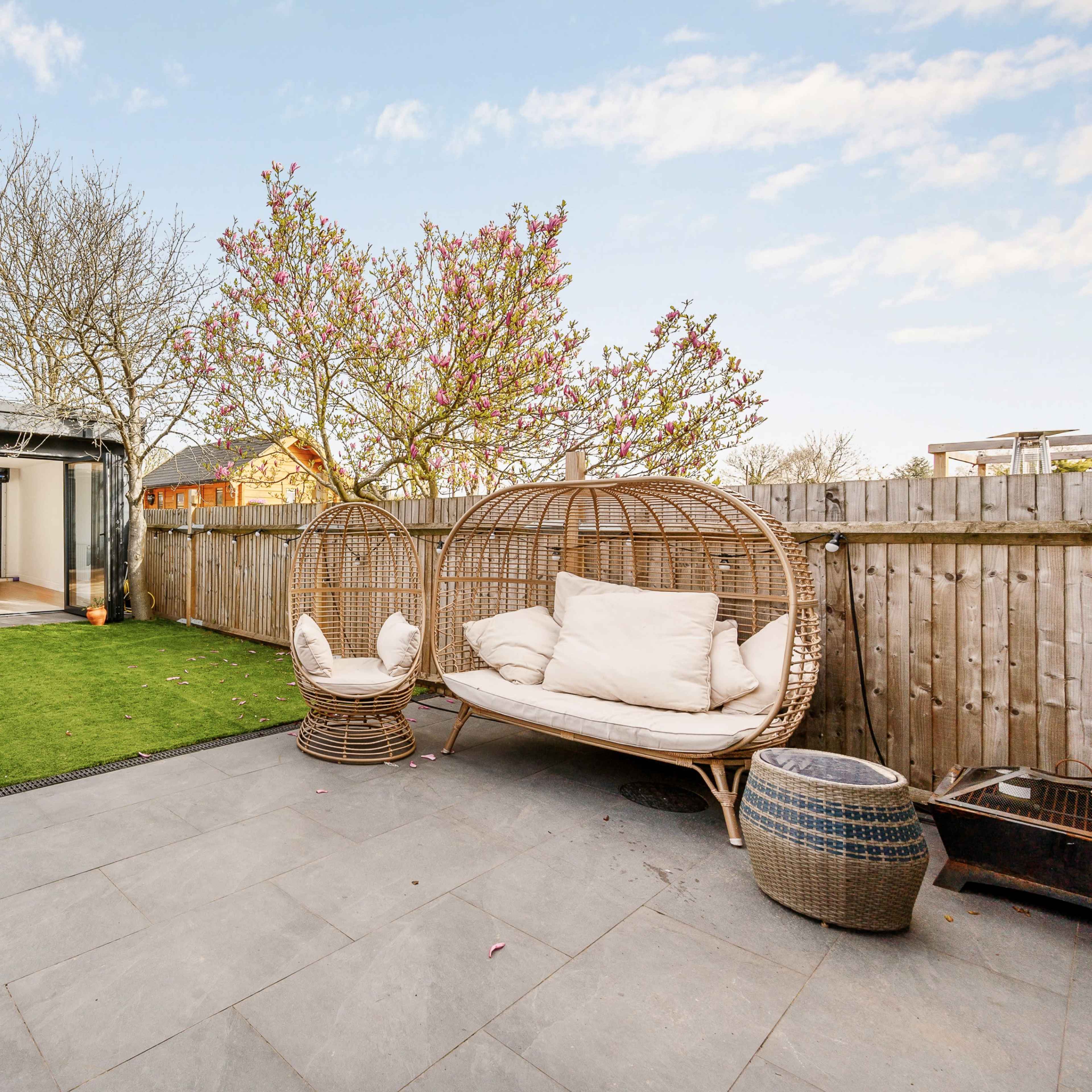 A landscaped outdoor area features two wicker chairs on a stone patio, surrounded by grass and a wooden fence, with a garage door and trees in the background.