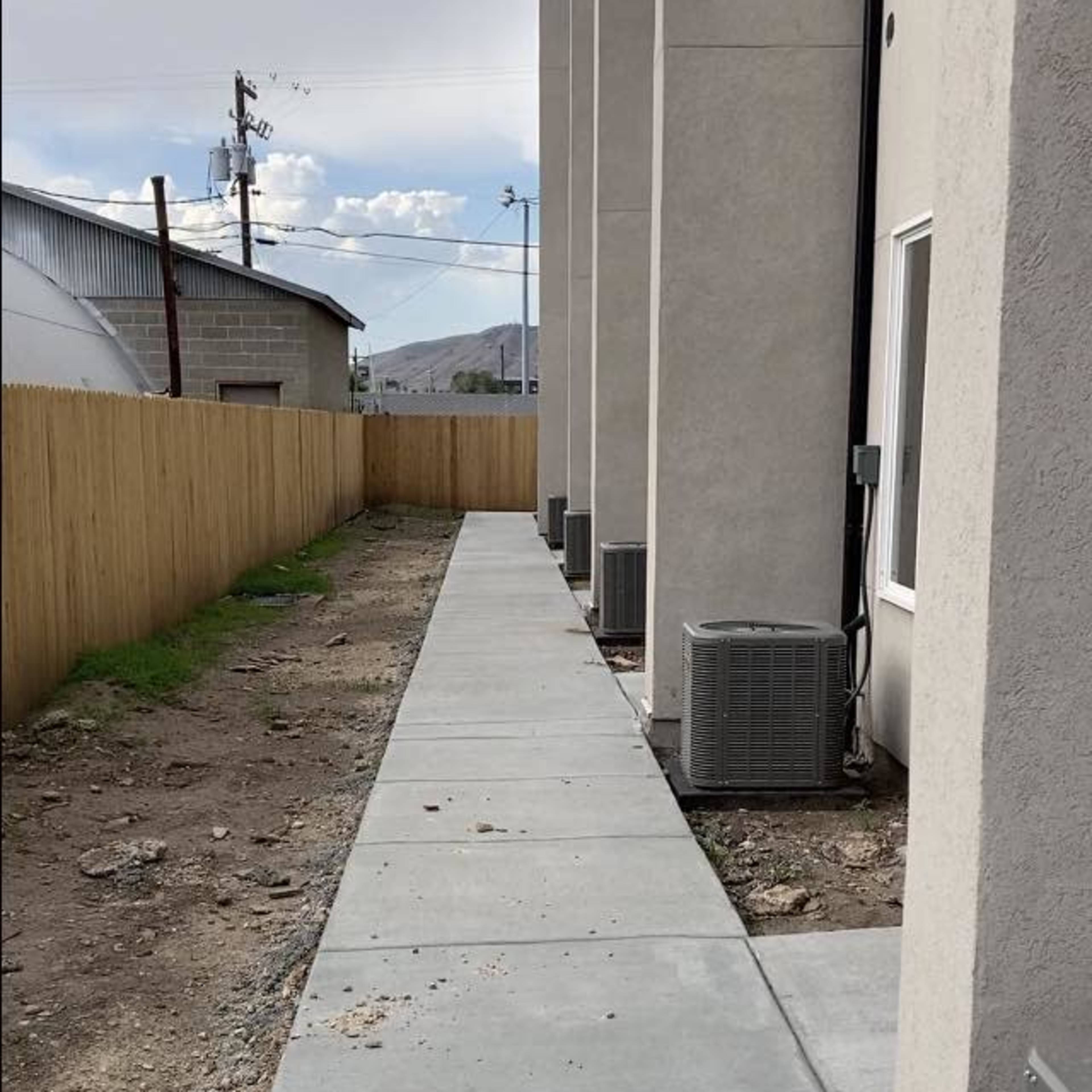 A concrete walkway runs alongside a building, bordered by a wooden fence and featuring air conditioning units.