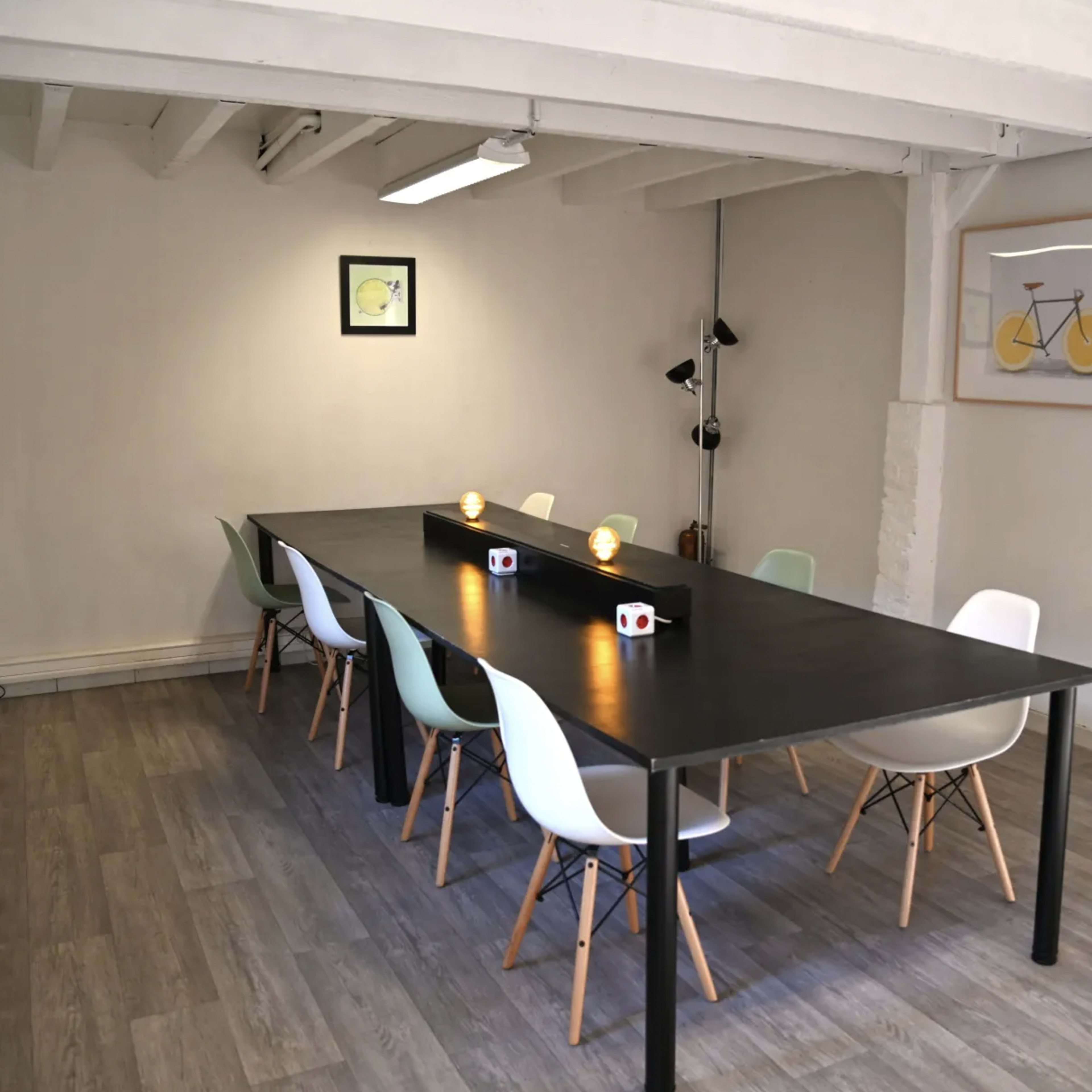 A long black table with chairs of various colors is set up in a well-lit, minimalistic room.
