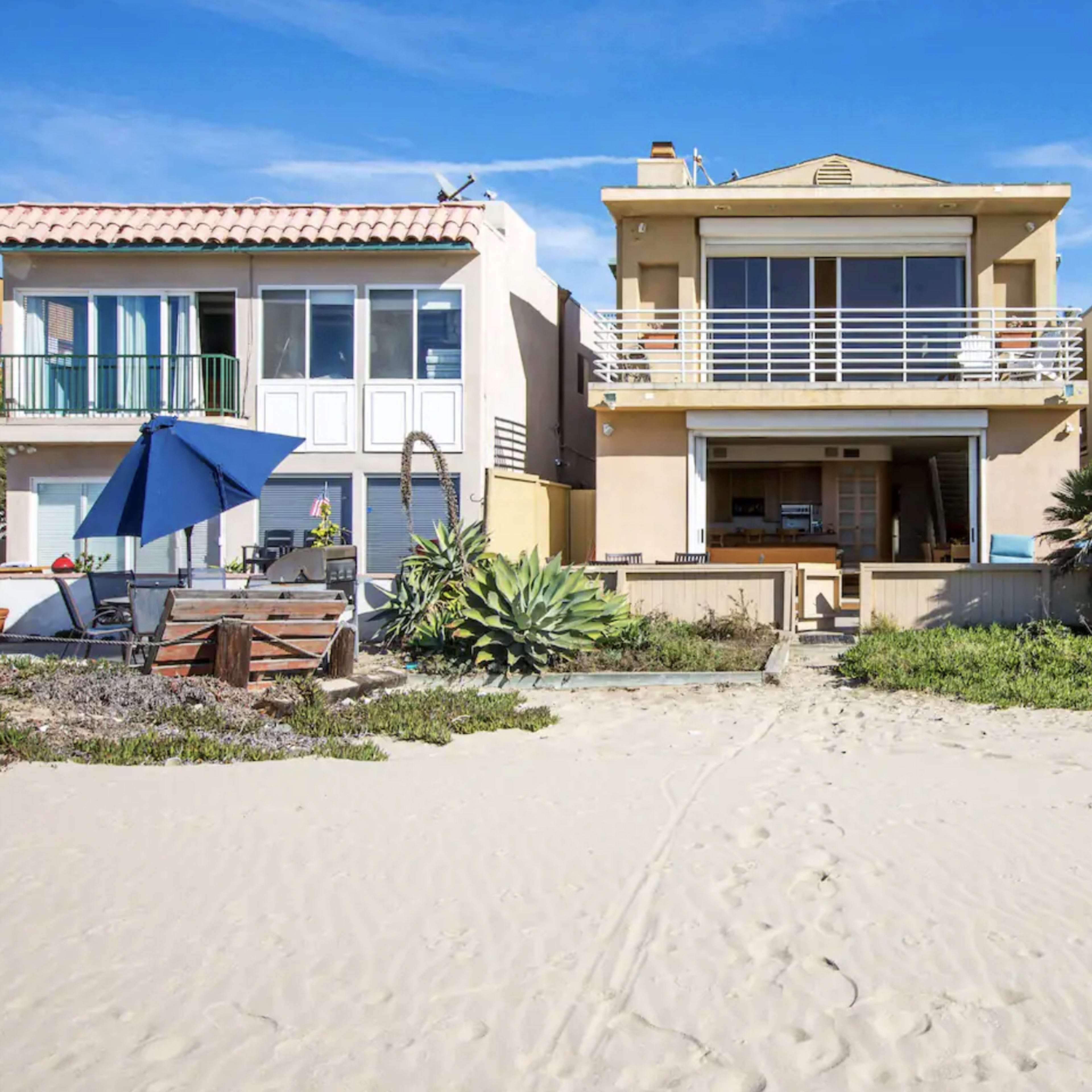 A beachfront property flanked by two neighboring houses, with sand leading up to the entrance.