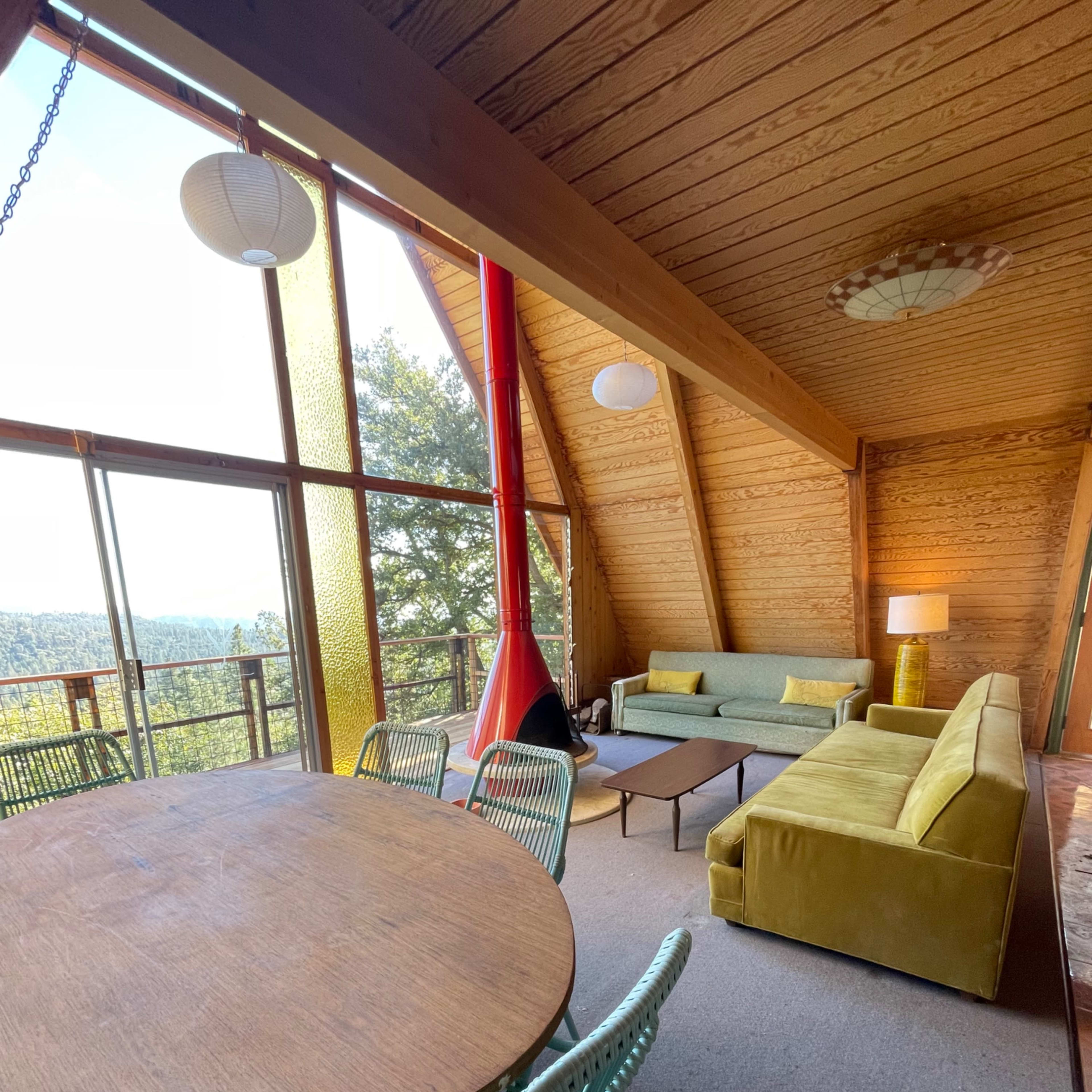 A modern living space with large windows offering a view of the outdoors, featuring a dining table with turquoise chairs, a green couch, and a yellow seating area.
