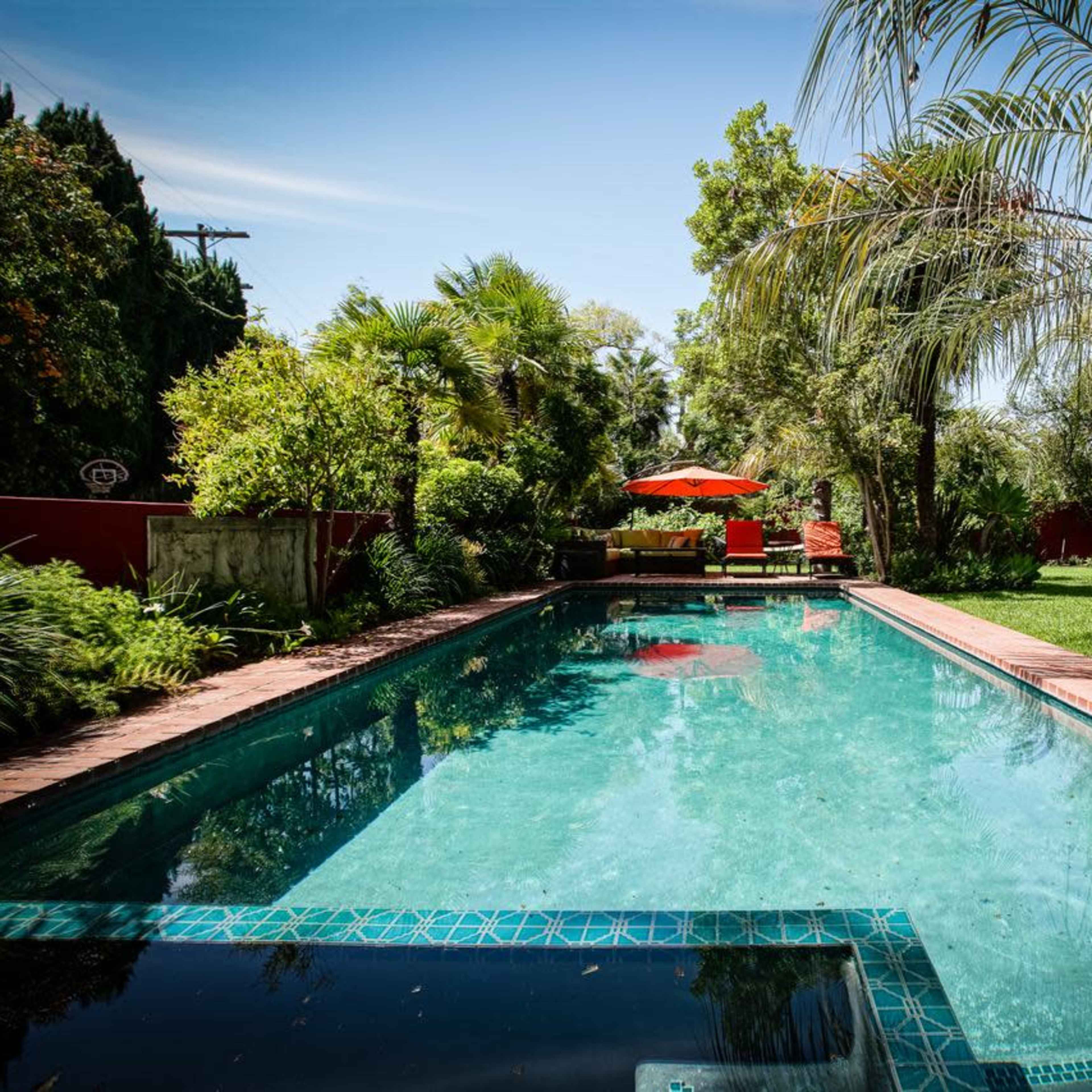A clear swimming pool is framed by lush greenery and a shaded seating area under an orange umbrella.