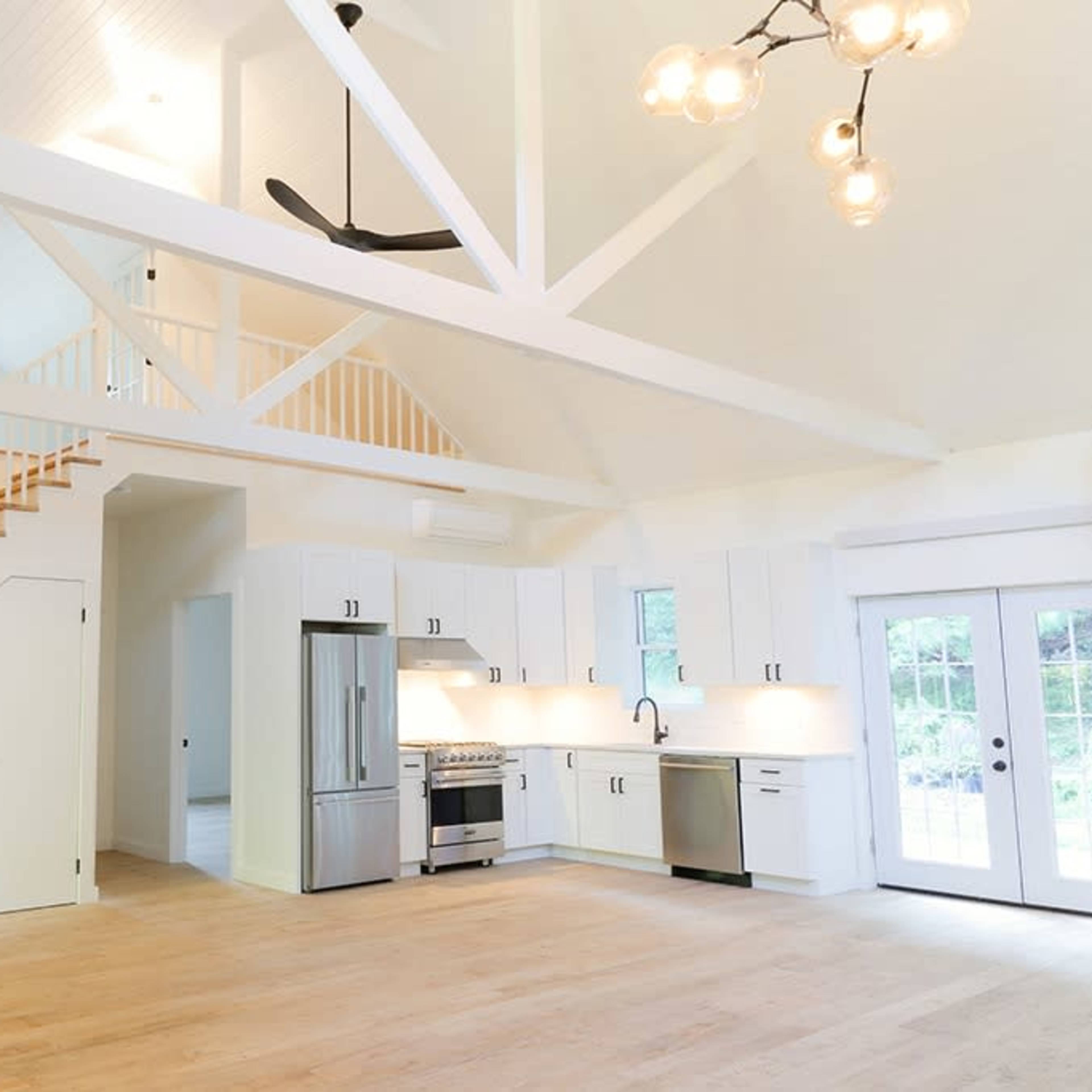 A bright, open-concept living area with a modern kitchen, a staircase leading to a loft, and large windows that allow natural light to enter.