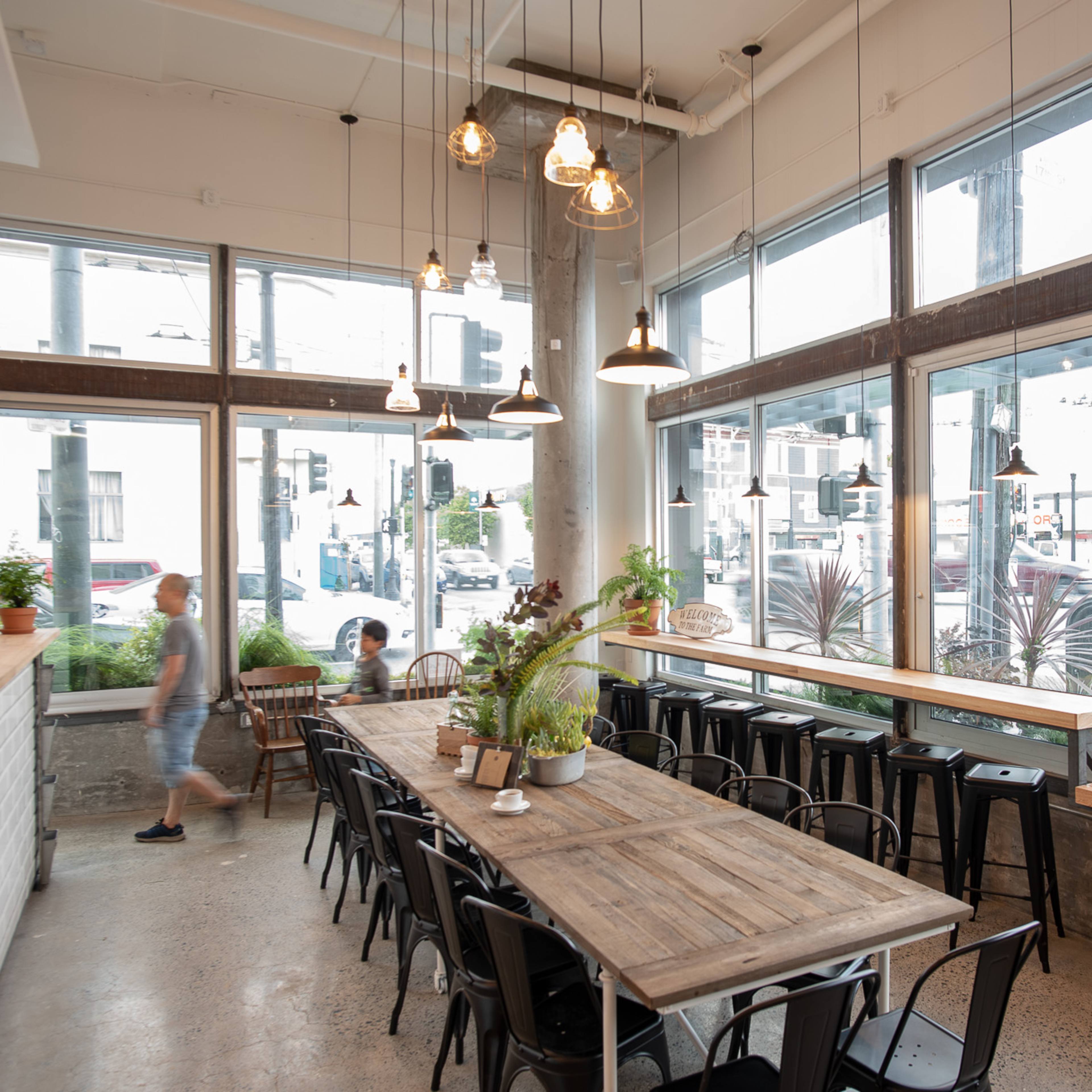 A spacious cafe features a large wooden table surrounded by black chairs, with large windows allowing natural light to fill the room.